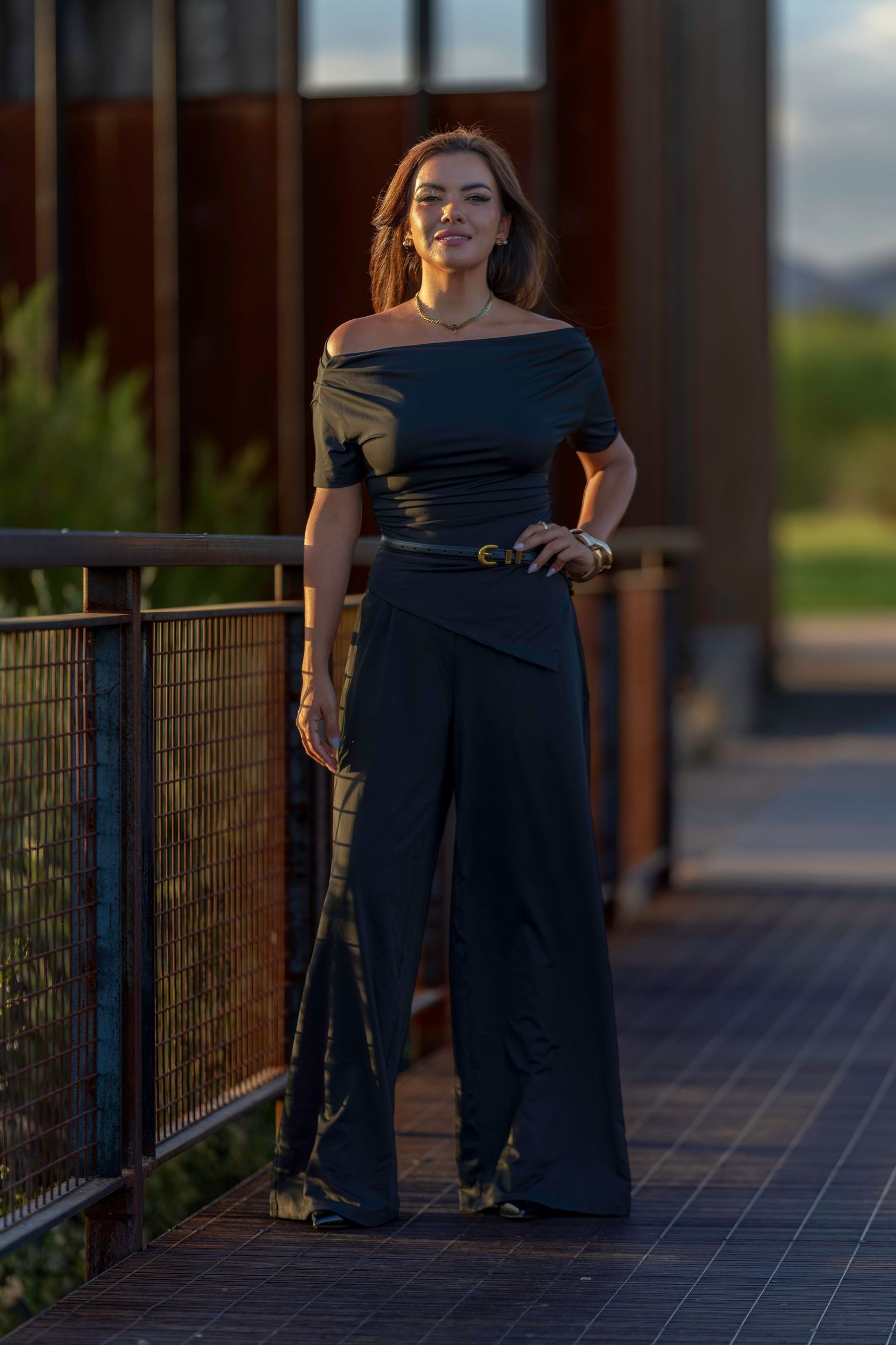 Woman in black off-shoulder top and wide-leg pants posing on outdoor walkway at golden hour.