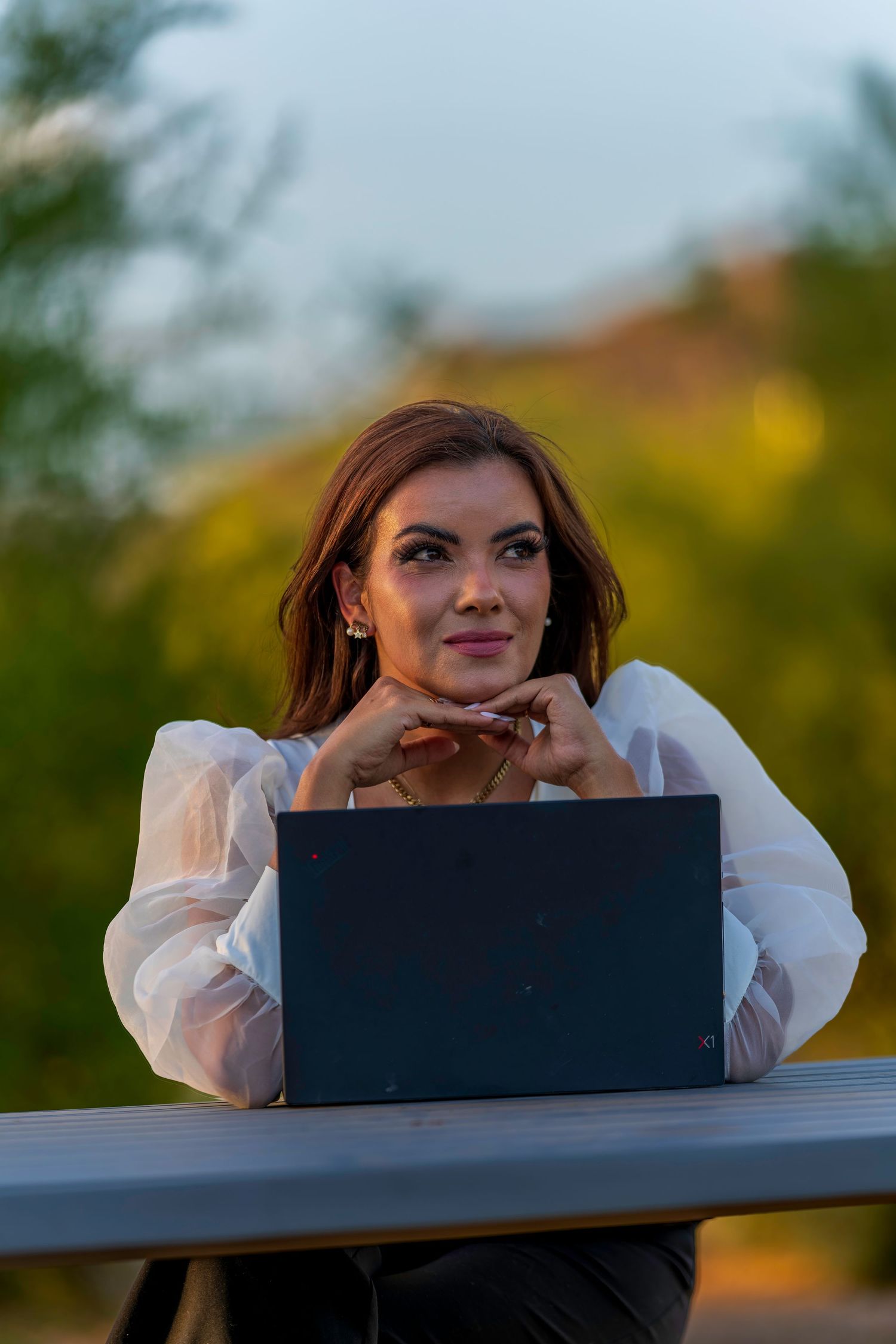 Woman in white blouse working on laptop outdoors with colorful autumn foliage background.