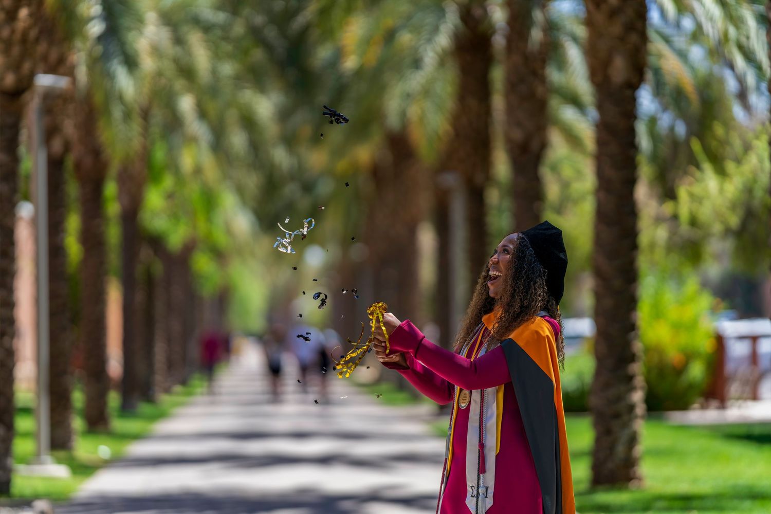 A woman in colorful graduation attire releases confetti outdoors along a palm tree-lined pathway on a sunny day.