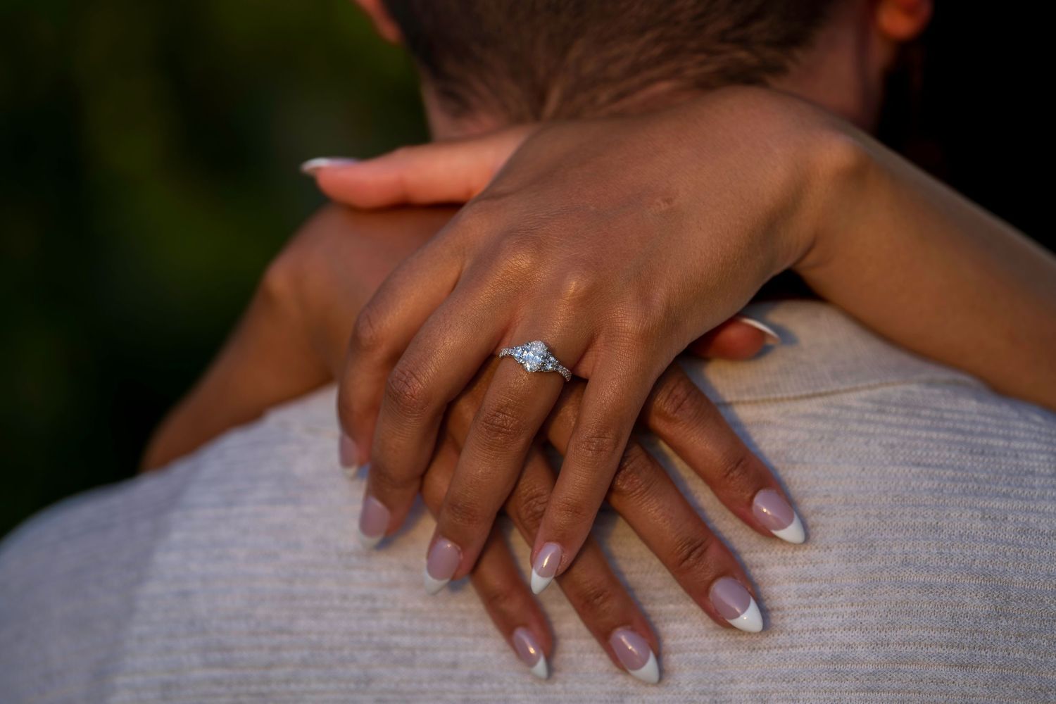 Close-up of a woman's hand with a diamond engagement ring resting on a man's shoulder in an outdoor setting.