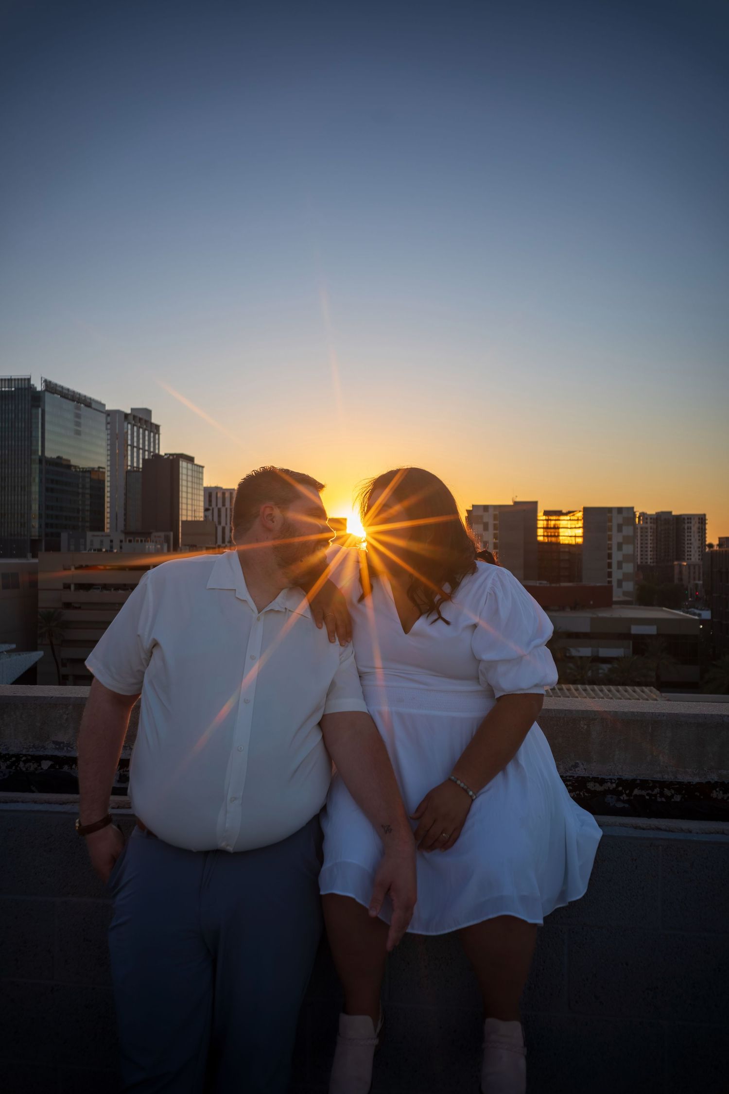 Couple holding hands at sunset on a rooftop with city skyline, sun flare between them, wearing white outfits.
