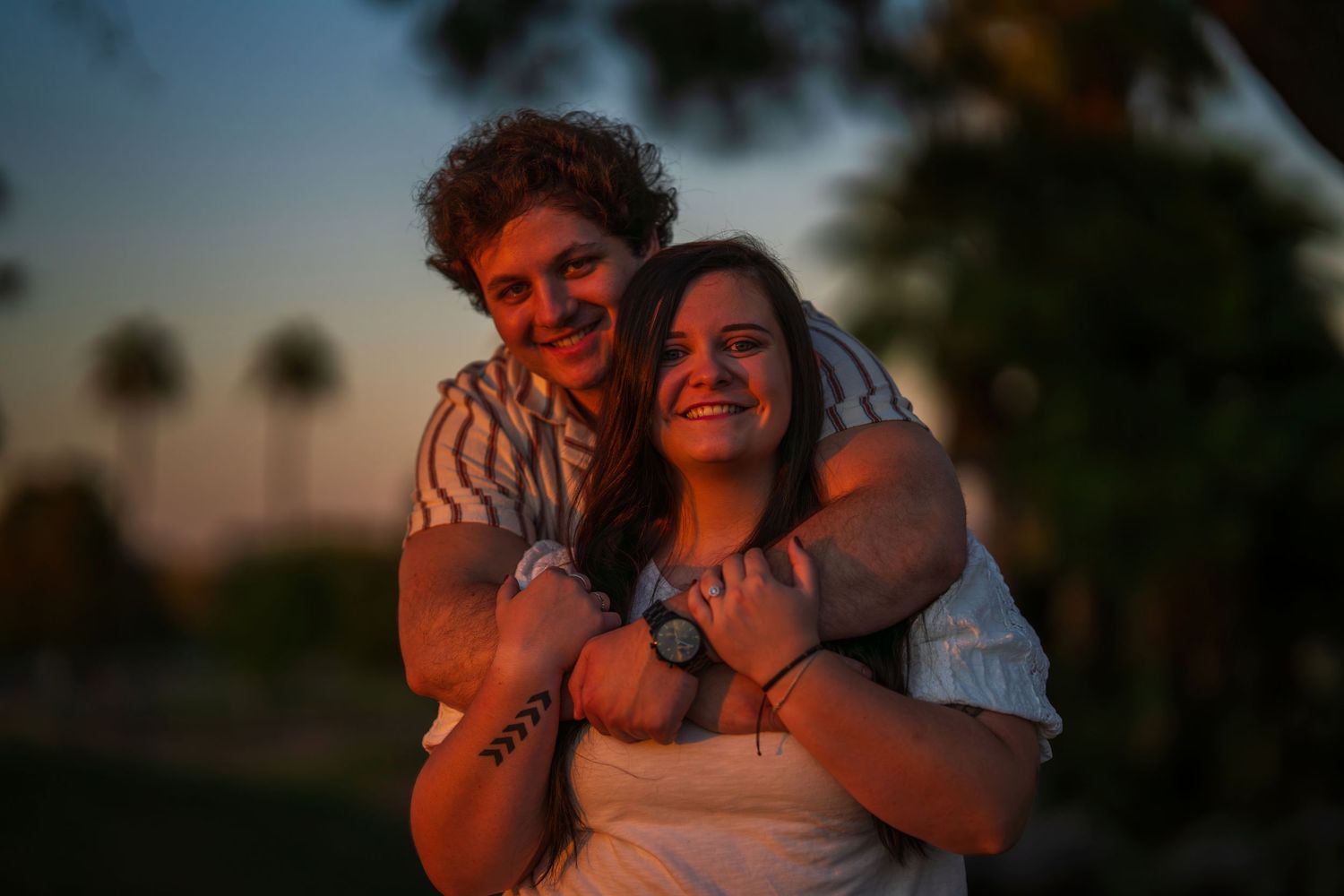 Happy couple embracing at sunset in park, man hugging woman from behind, both smiling warmly.