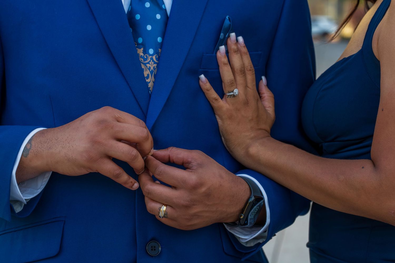 Close-up of couple's hands adjusting blue suit jacket, showcasing engagement ring and wedding band.