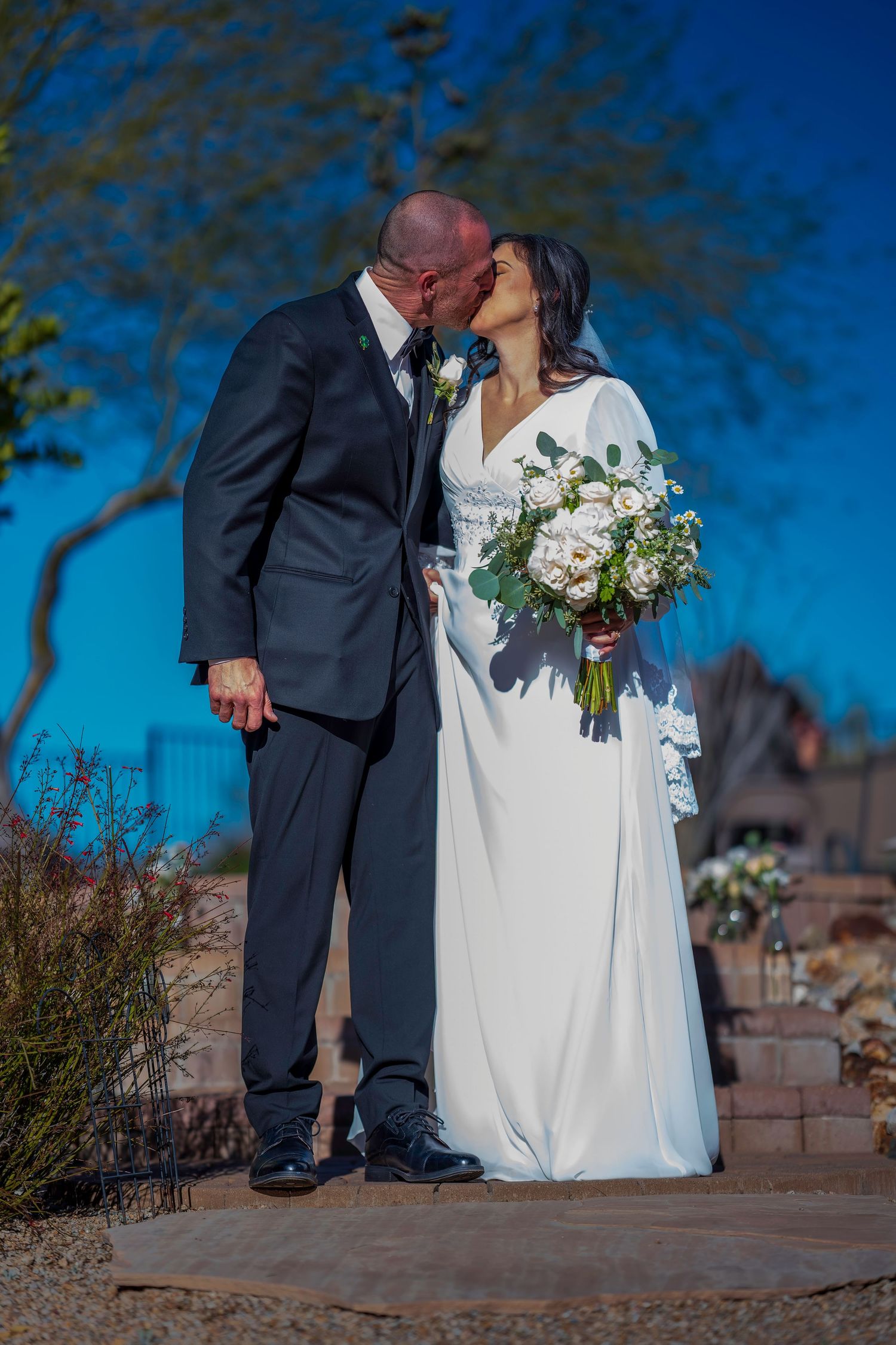 Newlyweds share a kiss outdoors, bride in white gown holding white bouquet under a bright blue sky.