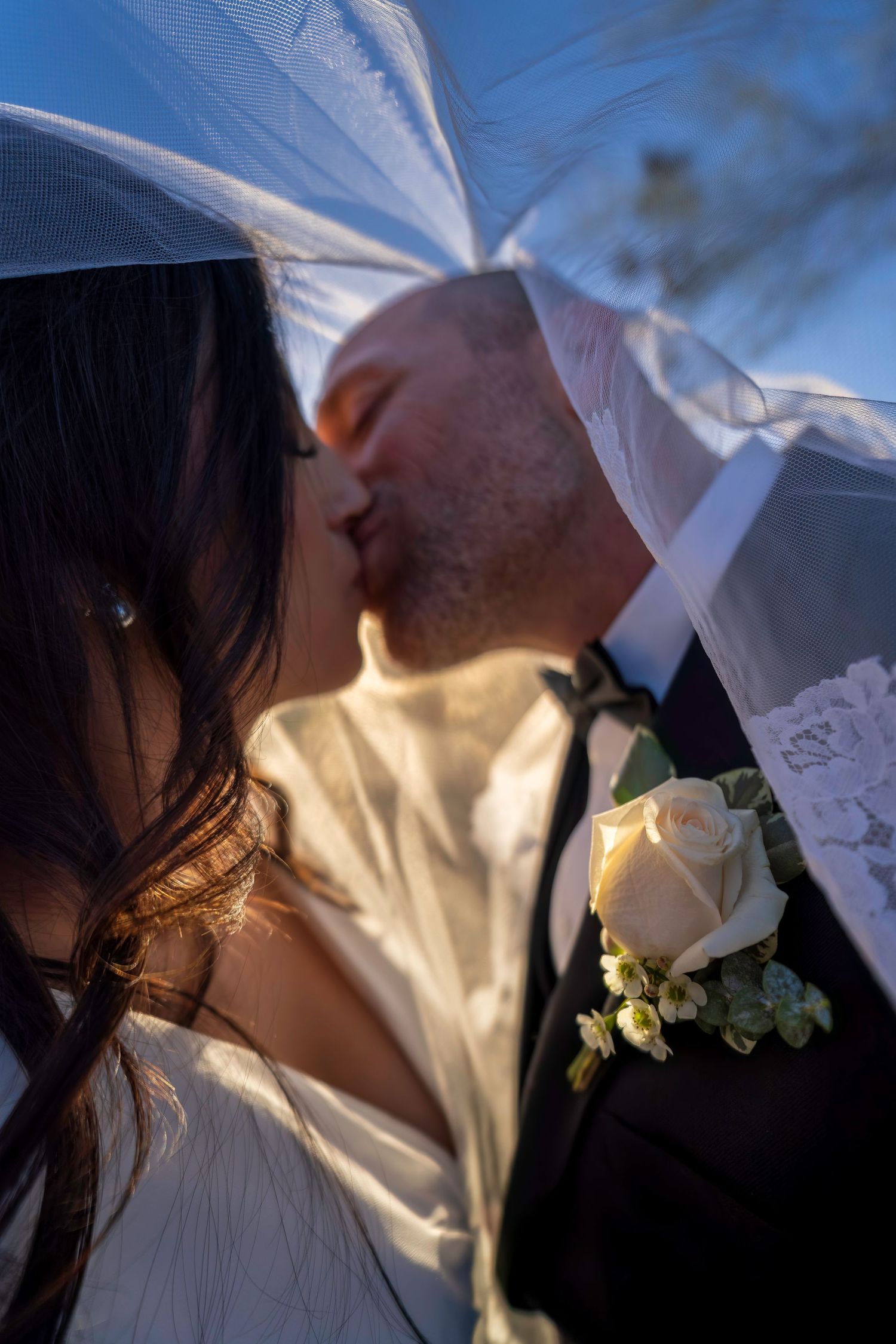 Bride and groom sharing romantic kiss under flowing white veil in soft golden sunlight.