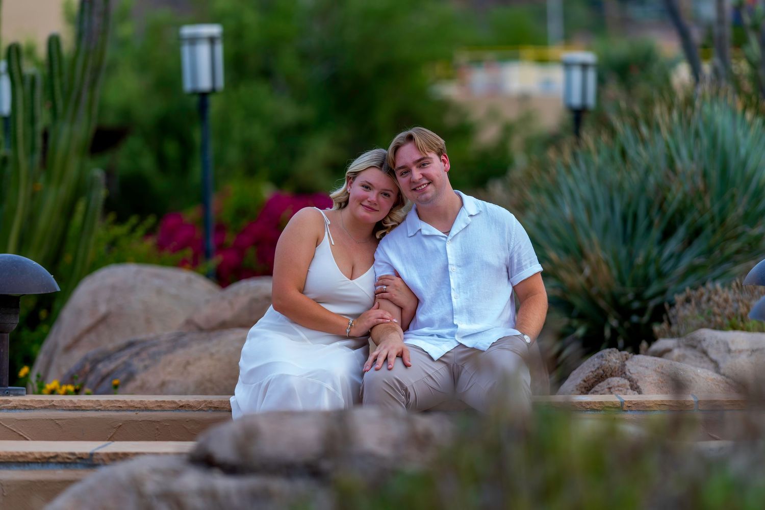 A smiling couple in white sit together on rocks surrounded by desert landscaping at dusk.
