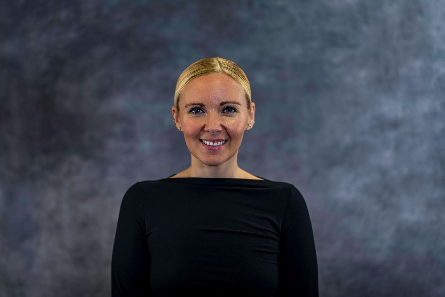 Professional headshot of someone wearing a black top against a dark textured background.