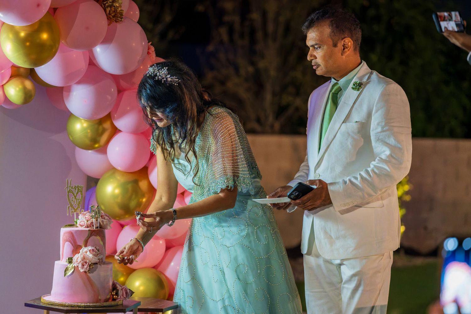 Couple cutting their wedding cake decorated with pink and gold balloons at night.