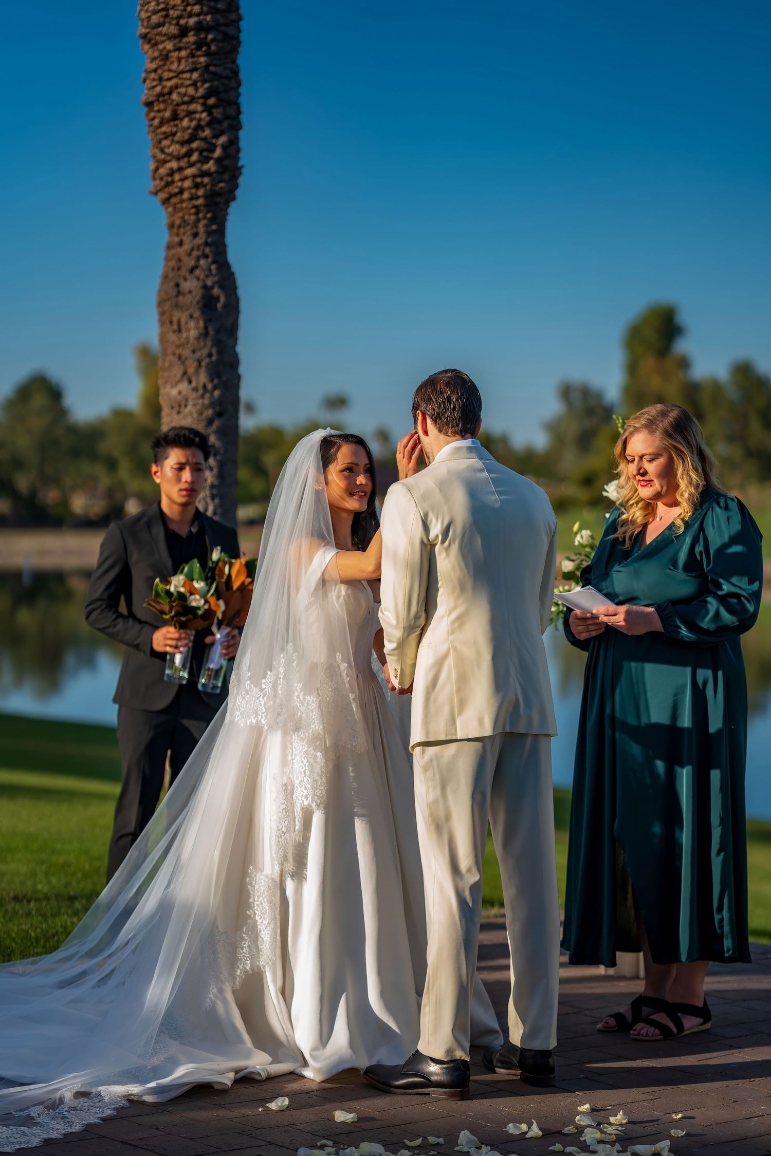 An outdoor lakeside wedding ceremony with palm trees and mountains in the background.