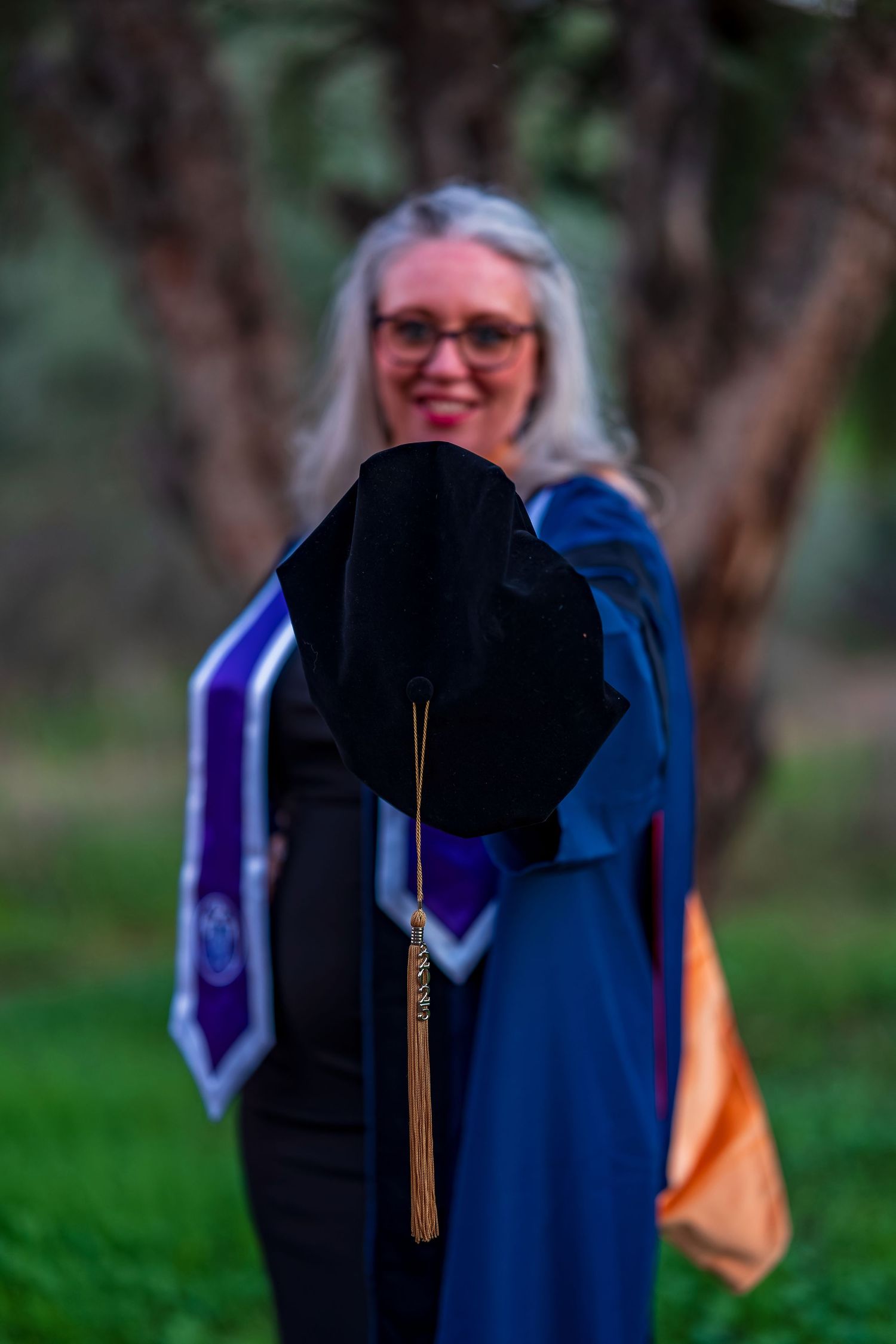 Graduation cap with gold tassel held up against blurred natural background.