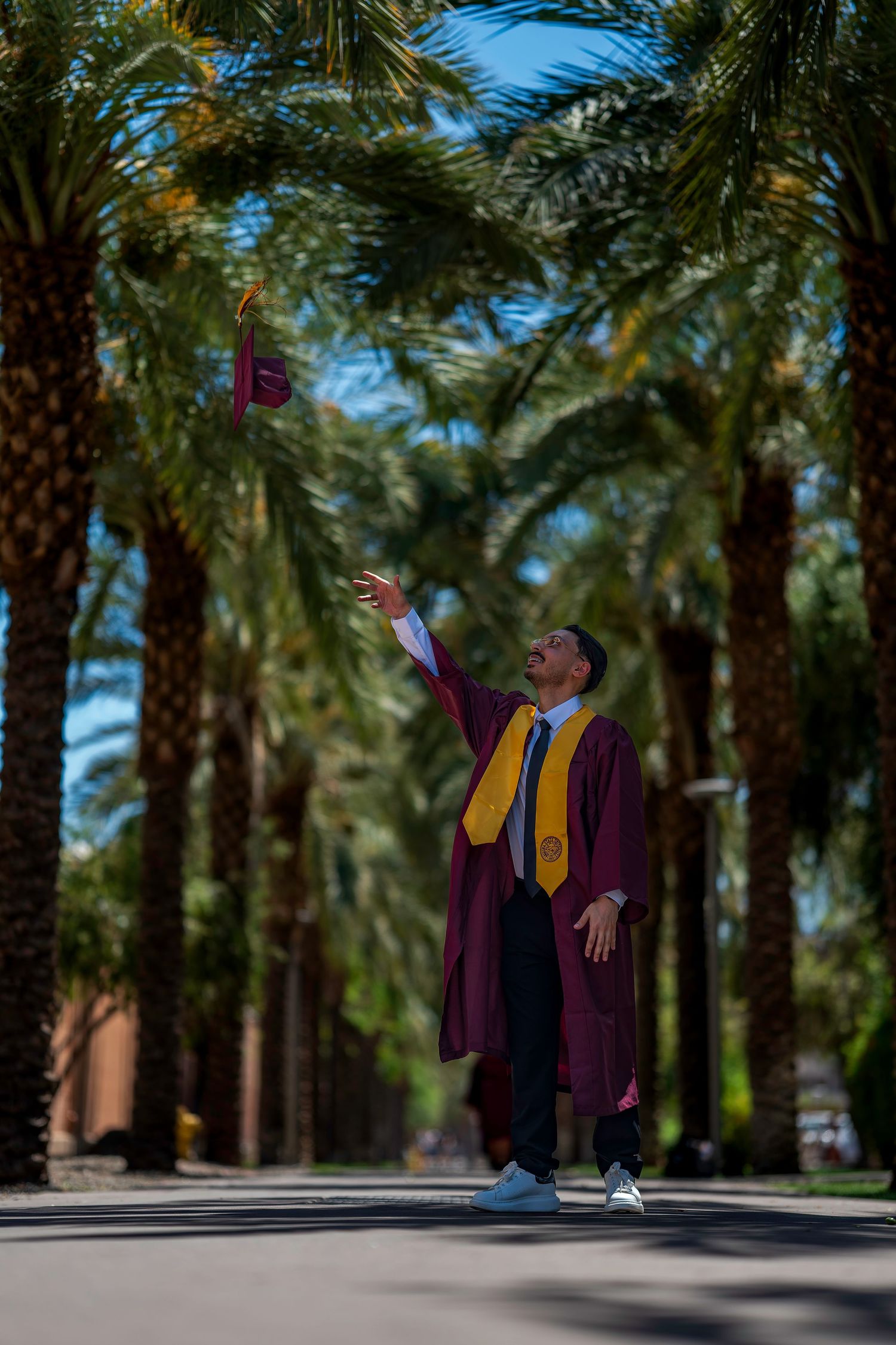 Graduate in maroon cap and gown tosses cap under tall palm trees on campus.