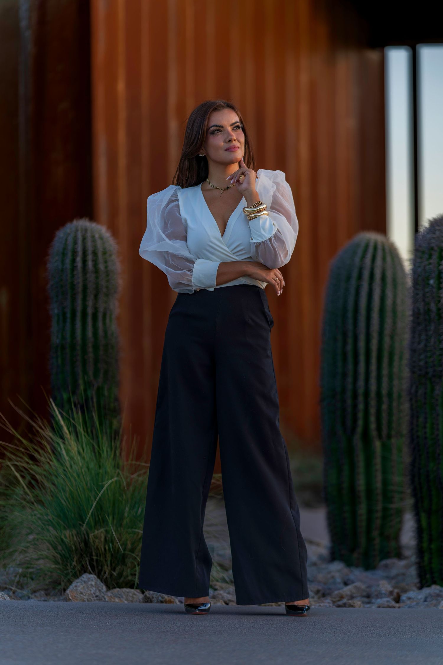 Stylish woman in white wrap blouse and wide-leg black pants posing near tall cacti.