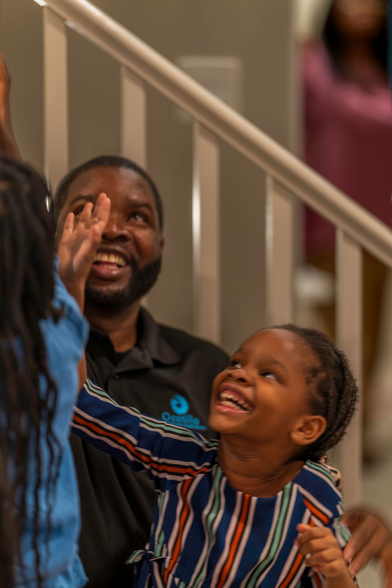 Man and young girl smiling and looking up joyfully near a staircase at a family event.