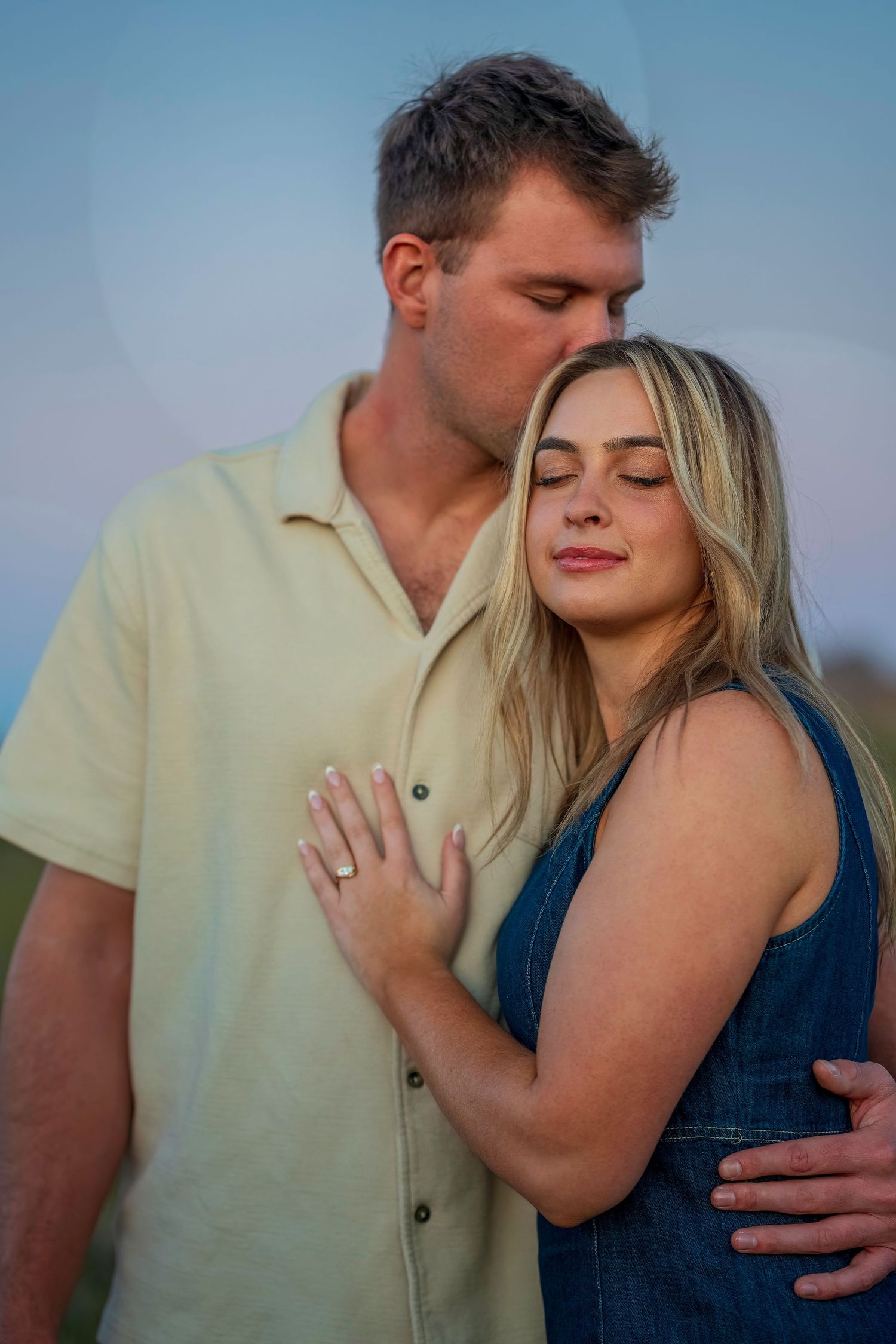 Romantic couple embrace outdoors, man kissing womans head, she displays engagement ring with eyes closed.
