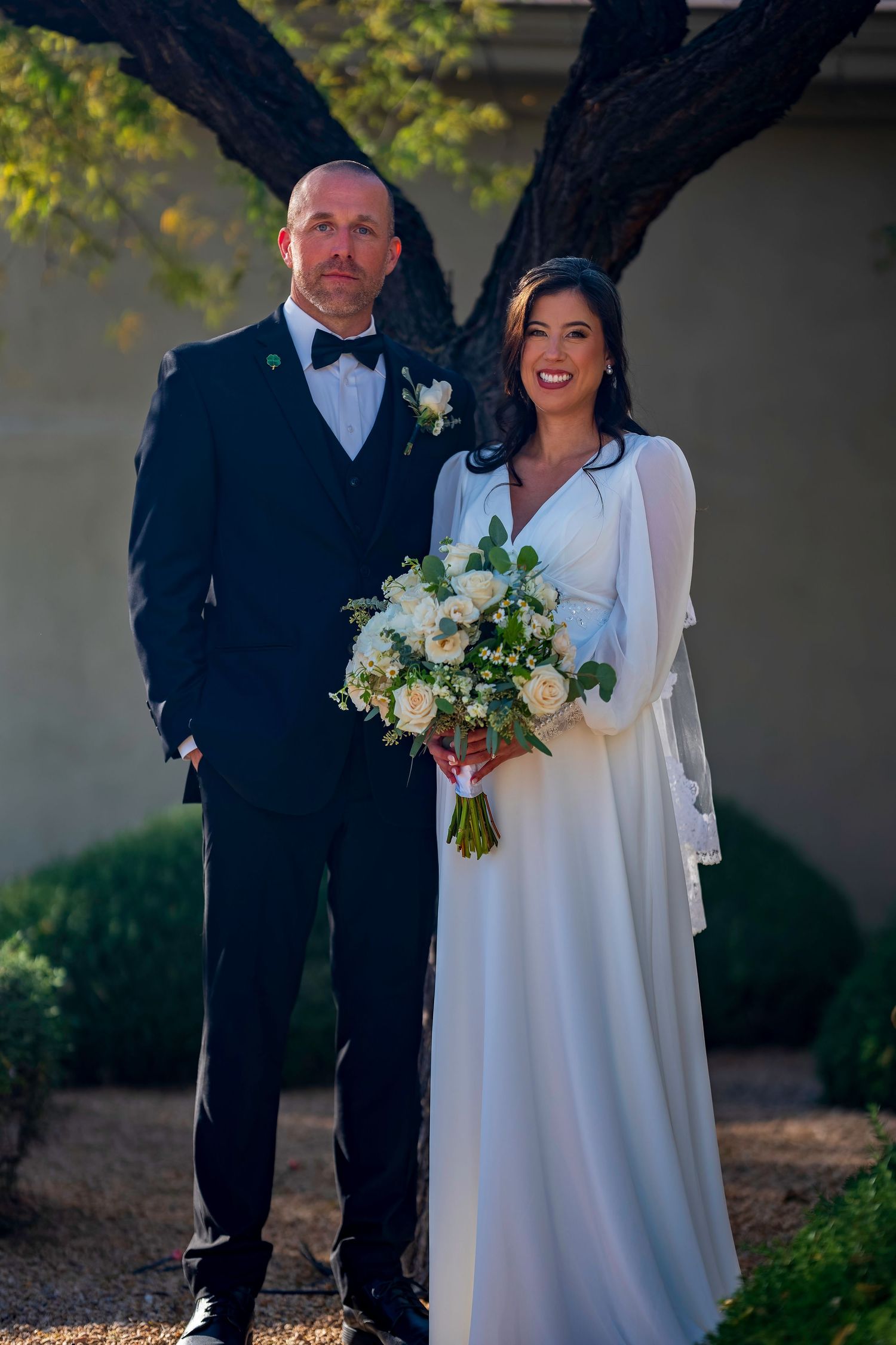 Bride and groom pose together outdoors, she holds white bouquet, he wears black tuxedo with boutonniere.