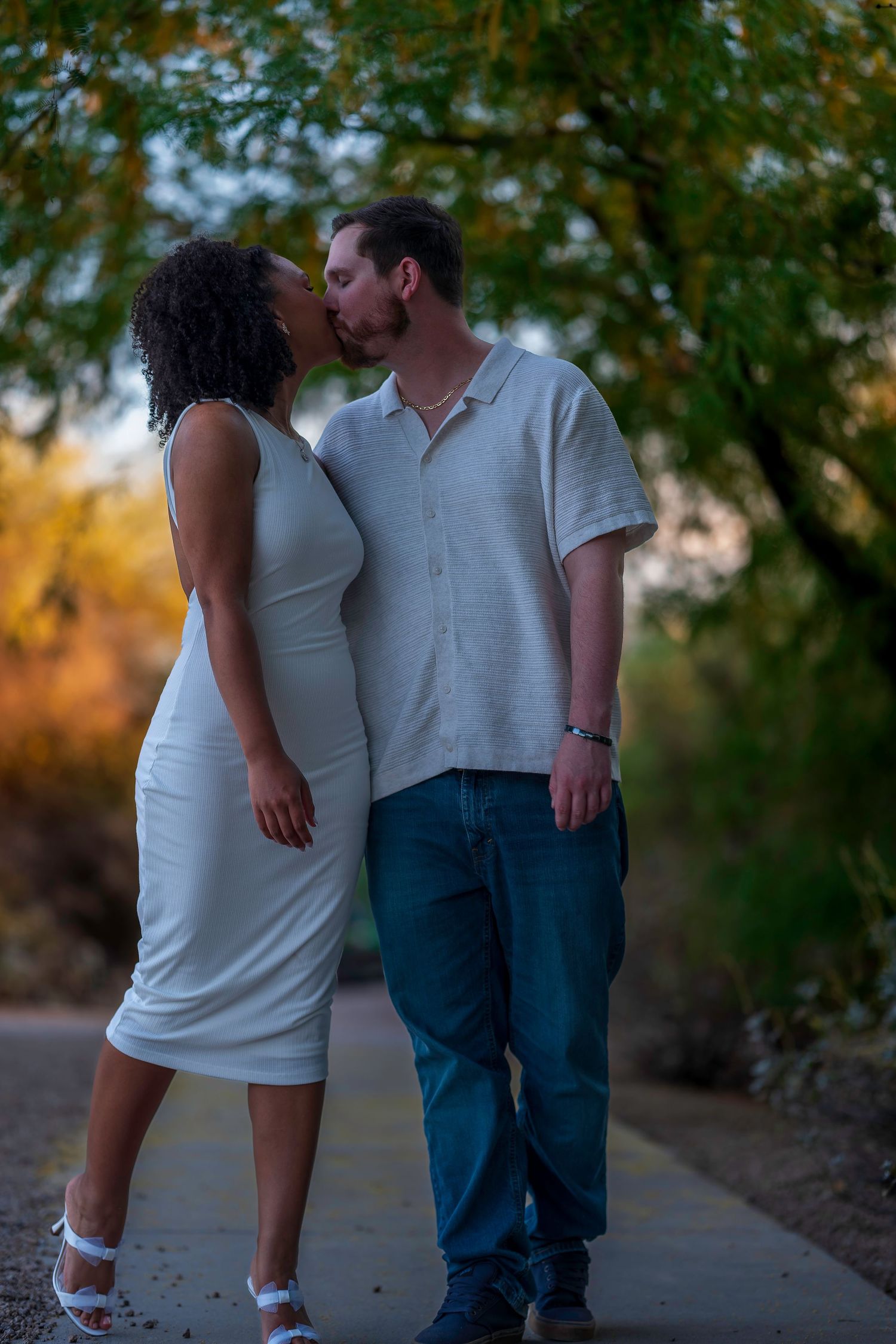 A couple sharing a romantic kiss on a scenic outdoor path, woman in white dress, man in jeans and white shirt.