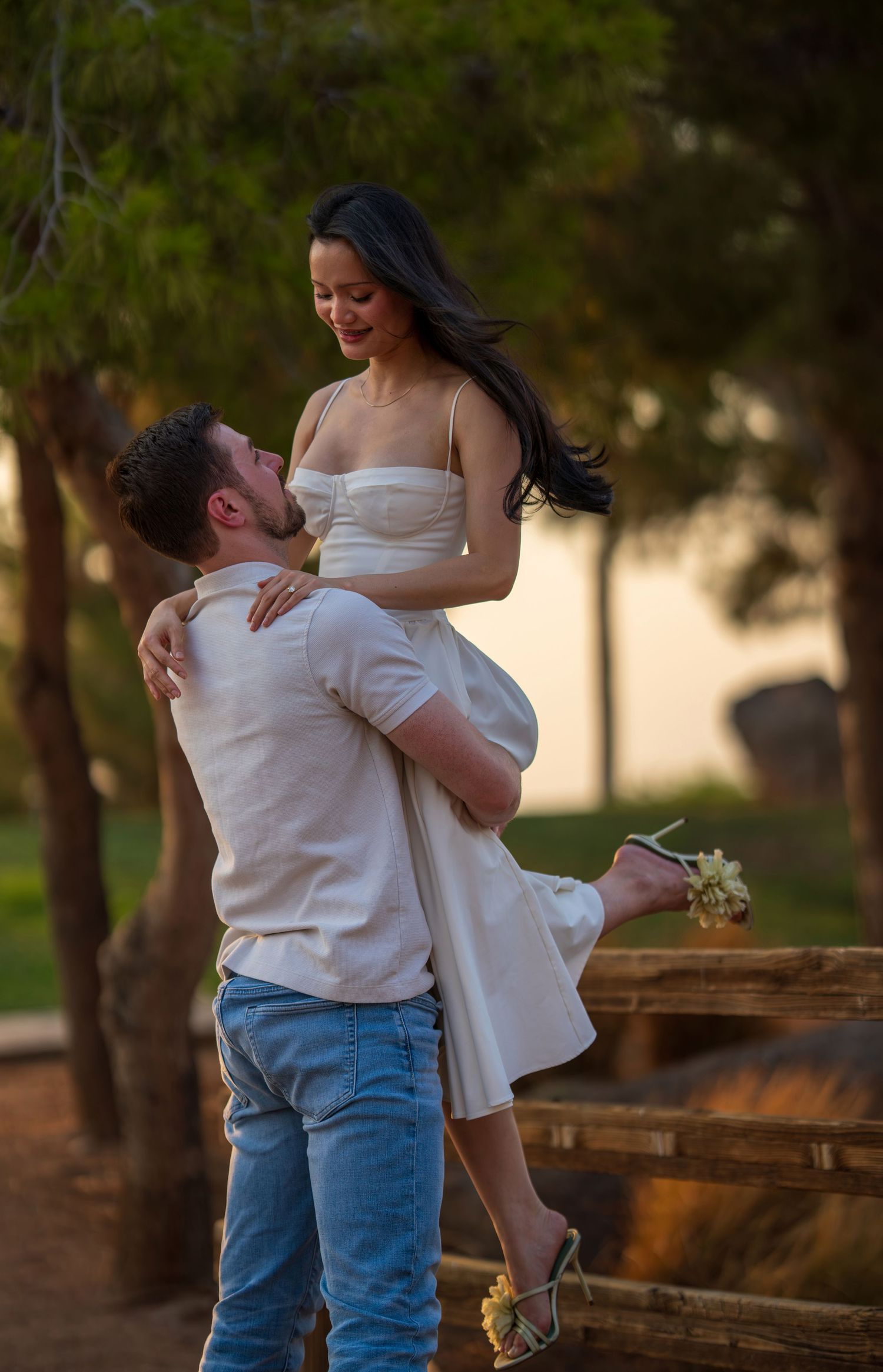 Man lifting woman in white strapless dress outdoors near trees during golden hour sunset.