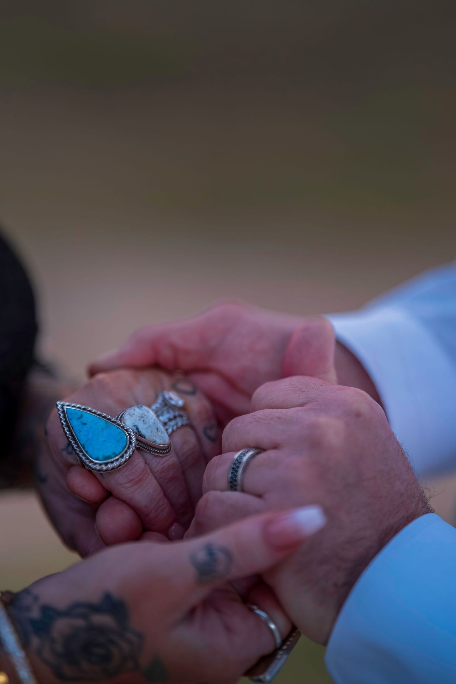 Close-up of two hands holding, adorned with turquoise rings, silver bands, and tattoos, symbolizing a romantic bond.