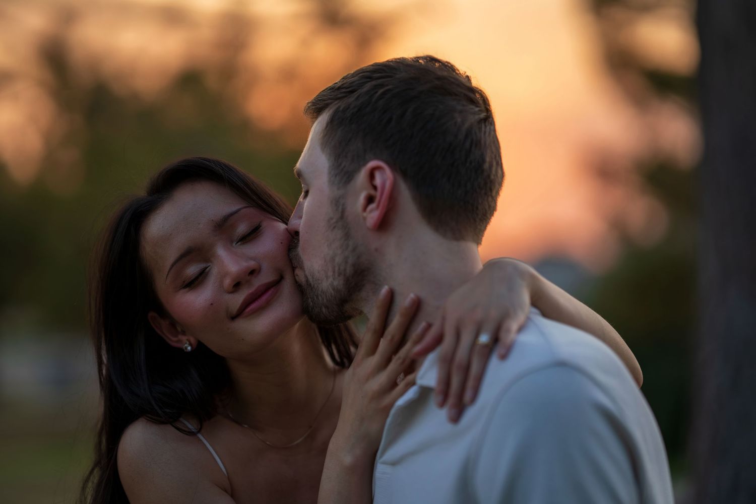 Couple sharing tender kiss at sunset, woman smiling as man kisses her cheek in golden light.