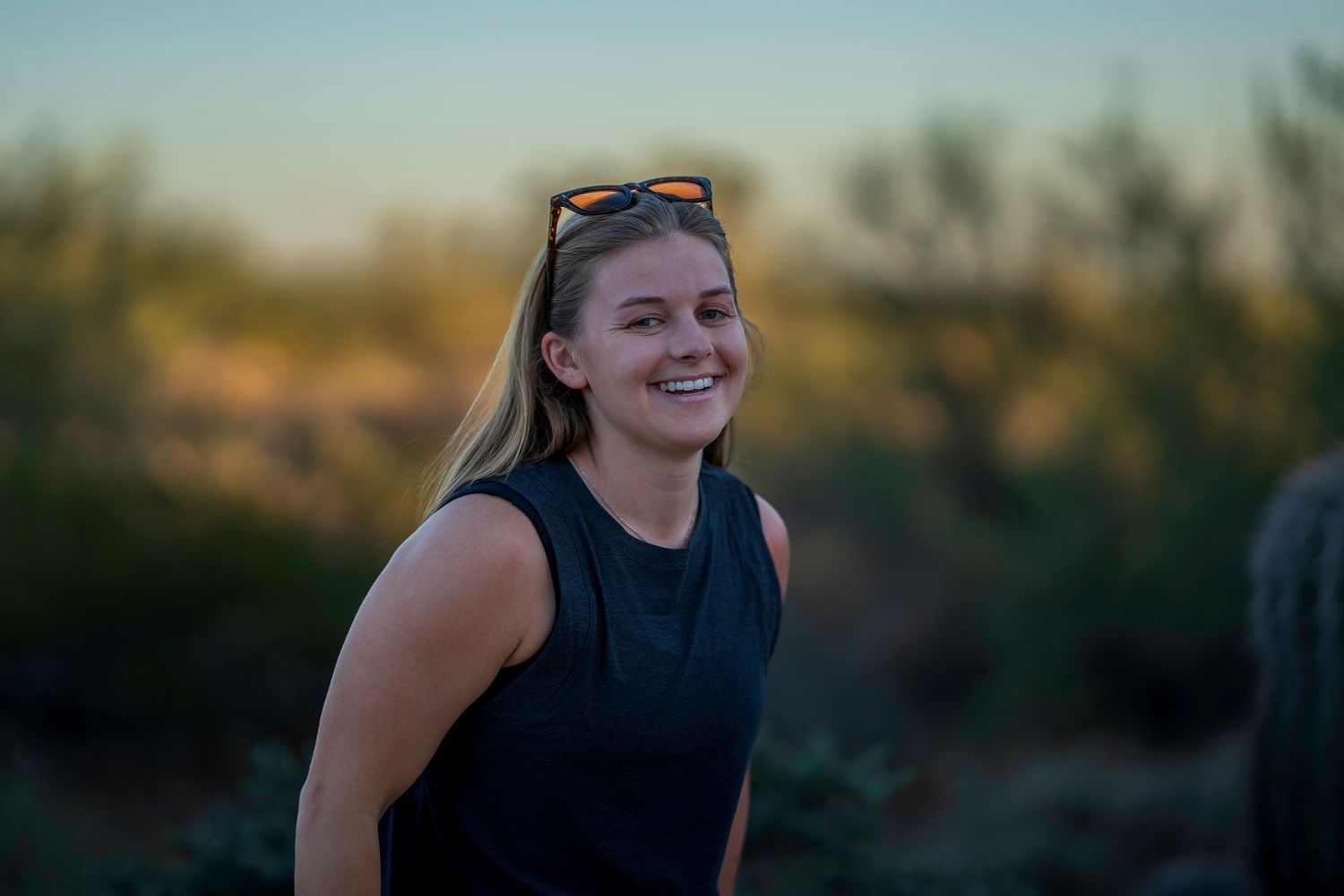 Smiling blonde woman with sunglasses on head wearing black sleeveless top outdoors at sunset in desert landscape.