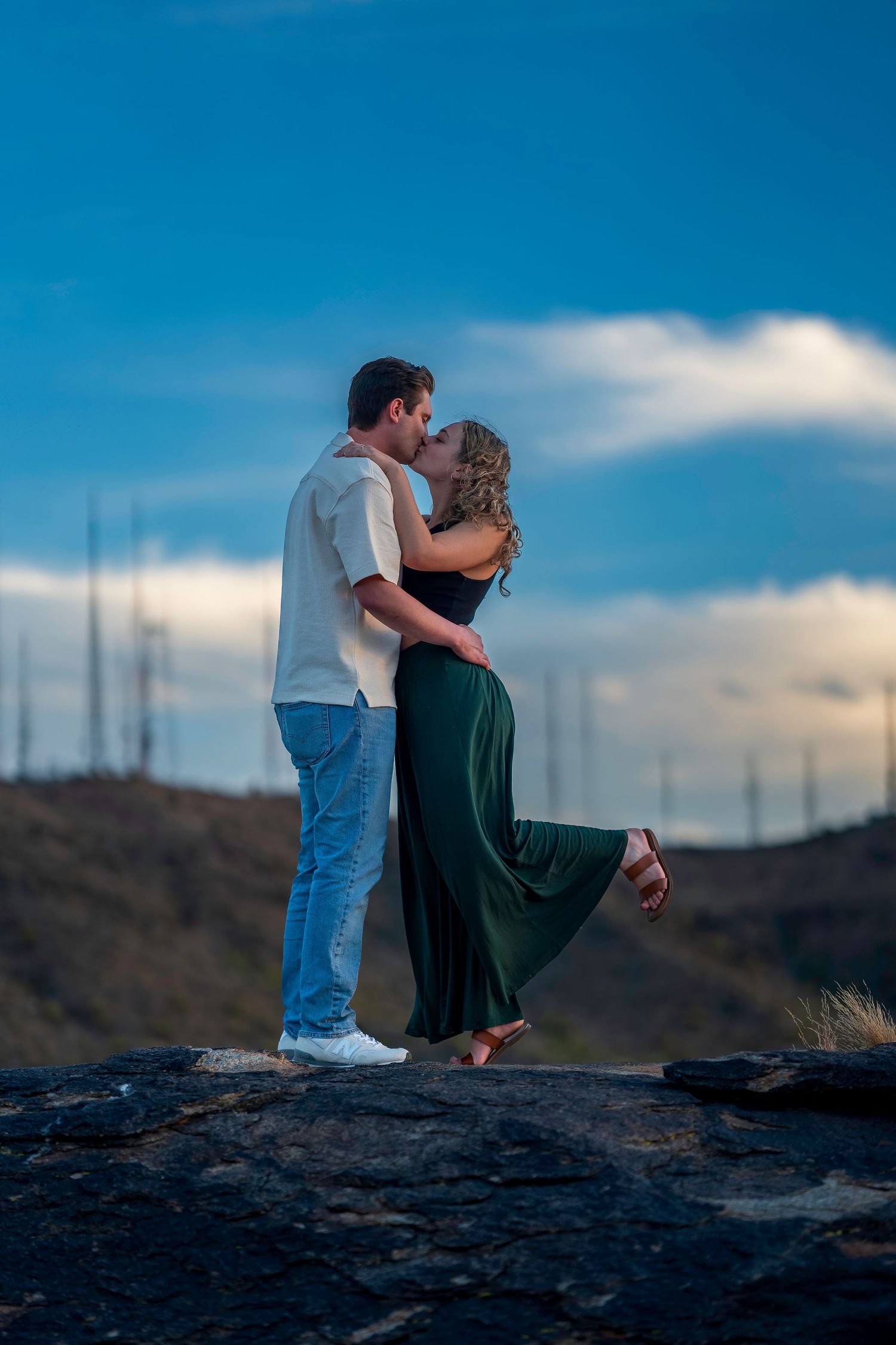 A romantic couple shares a kiss while standing on rocks with a scenic mountain backdrop at dusk.