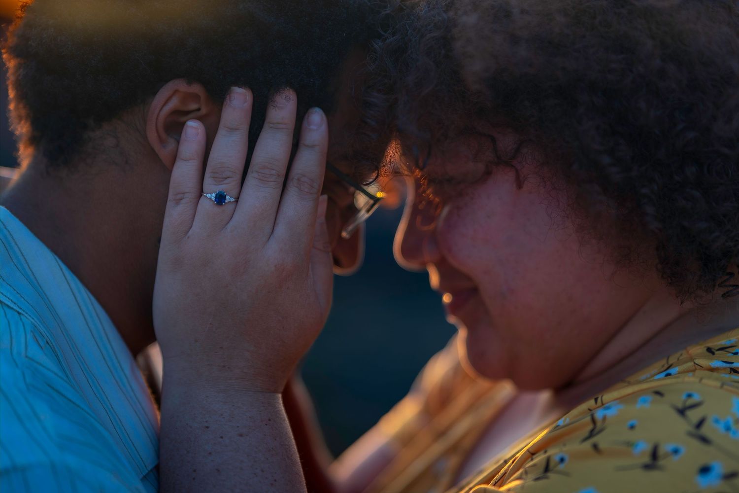 Two women share an intimate forehead-to-forehead moment, one wearing a blue ring, bathed in warm golden light.