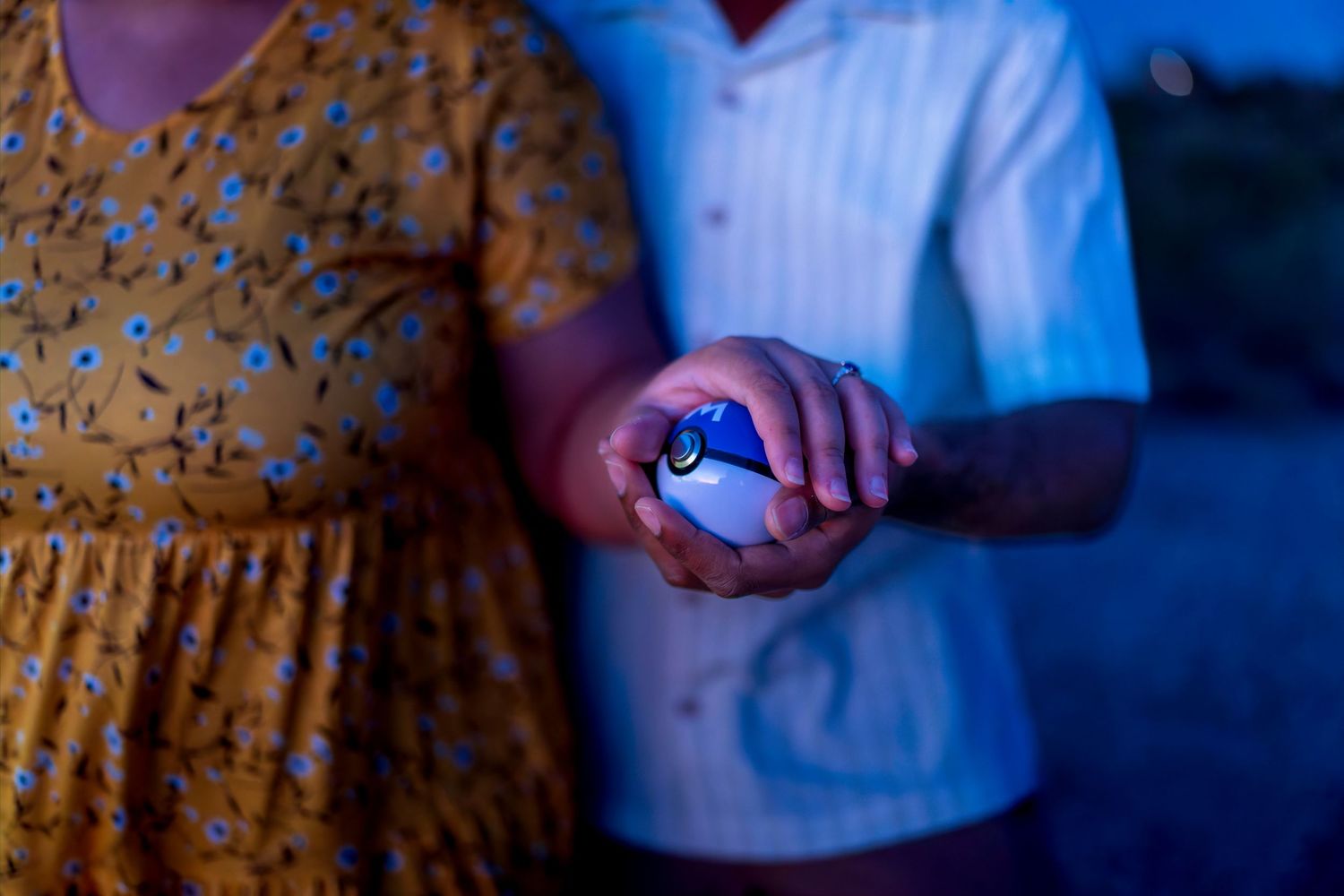 Couple holding illuminated blue Pokeball together during nighttime engagement photography session outdoors.