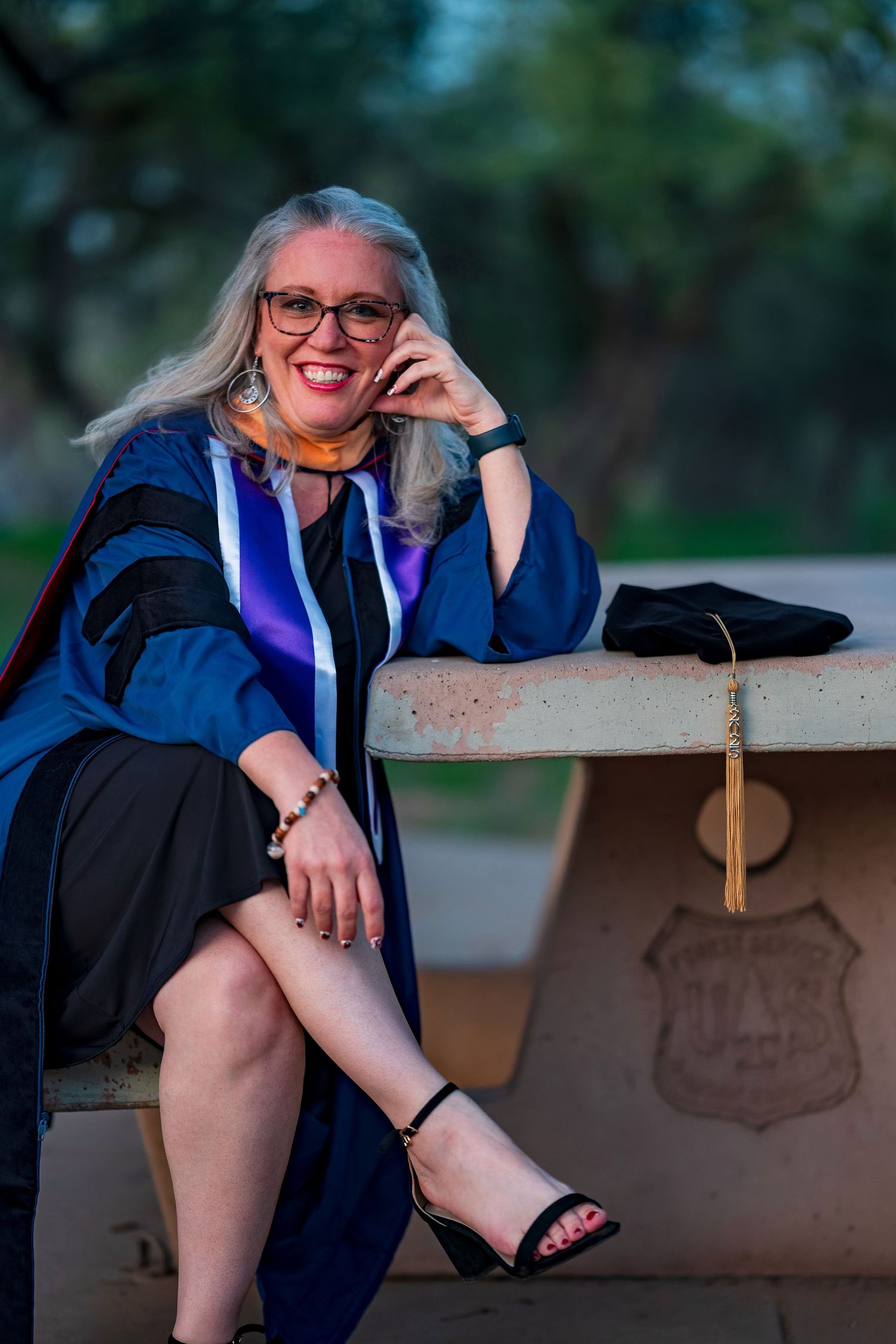 Graduate in academic regalia sits casually on bench during outdoor photo session.