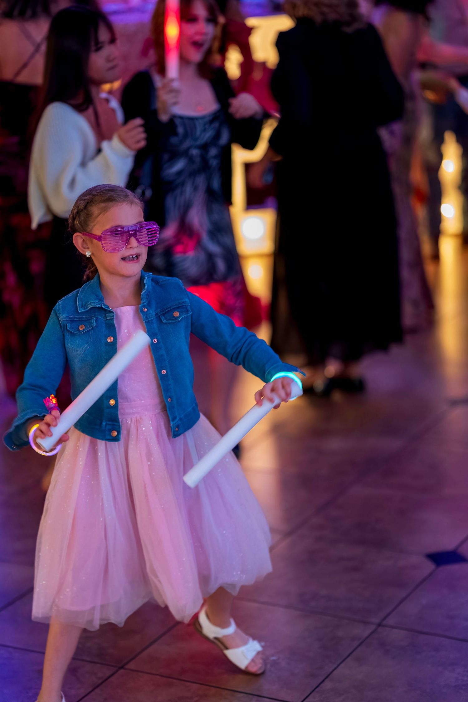 Young girl in pink tutu dress and denim jacket dancing with glow sticks at a party.