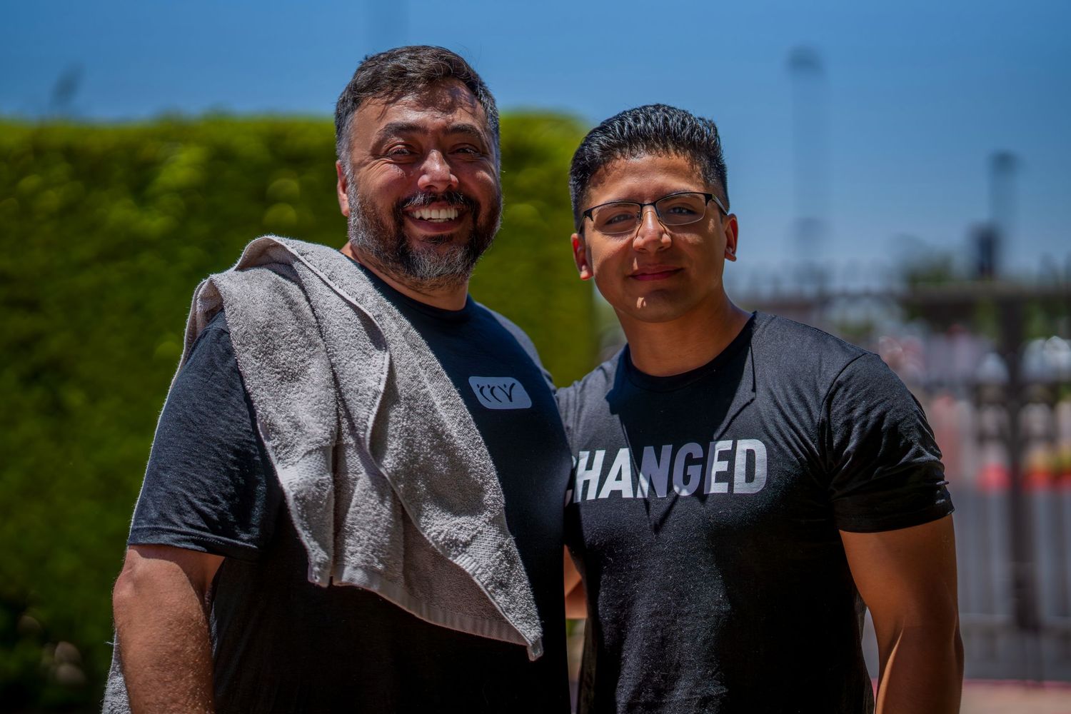 Two smiling men in casual t-shirts pose outdoors on a sunny day, one wearing a shirt that reads Changed.