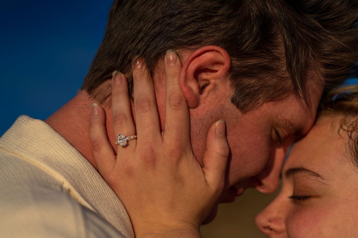 Close-up of couple embracing, woman wearing diamond engagement ring touching man's face against blue sky.