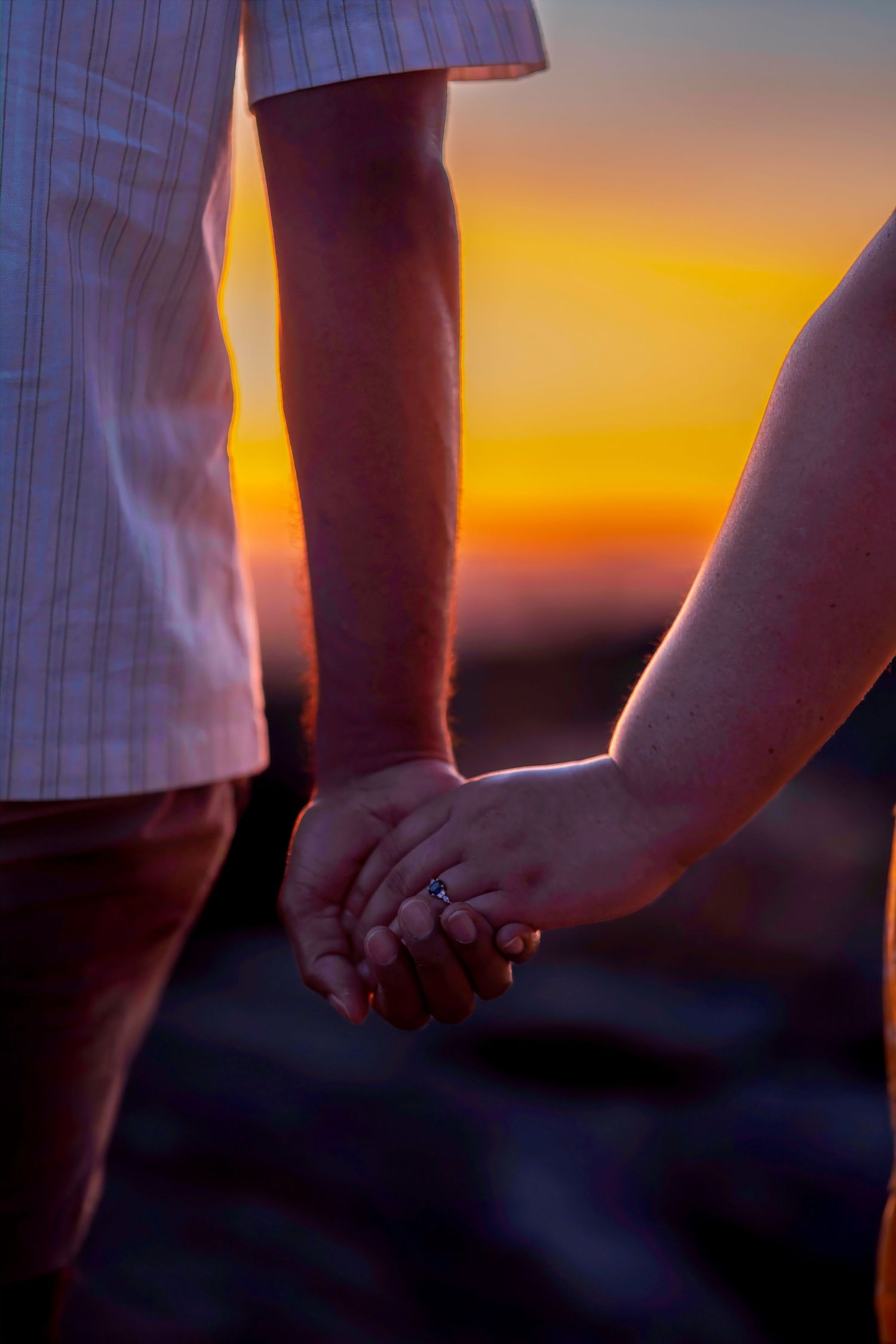 Couple holding hands at sunset with vivid orange sky, engagement ring visible on womans finger.