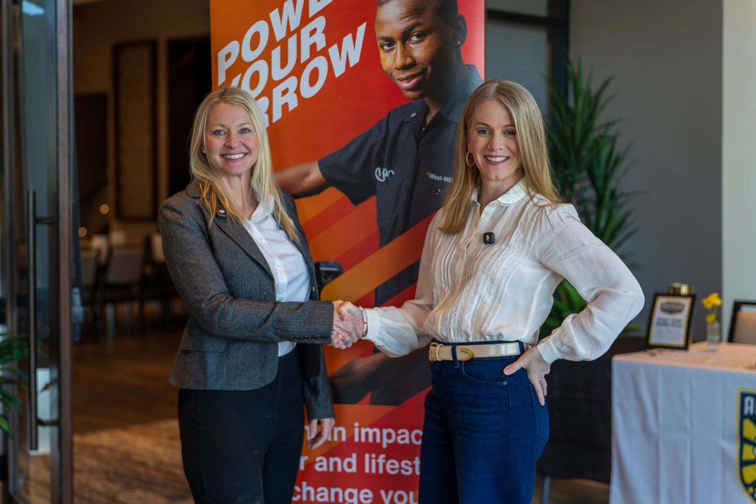 Two professional women shake hands in front of an orange motivational banner at a business networking event.
