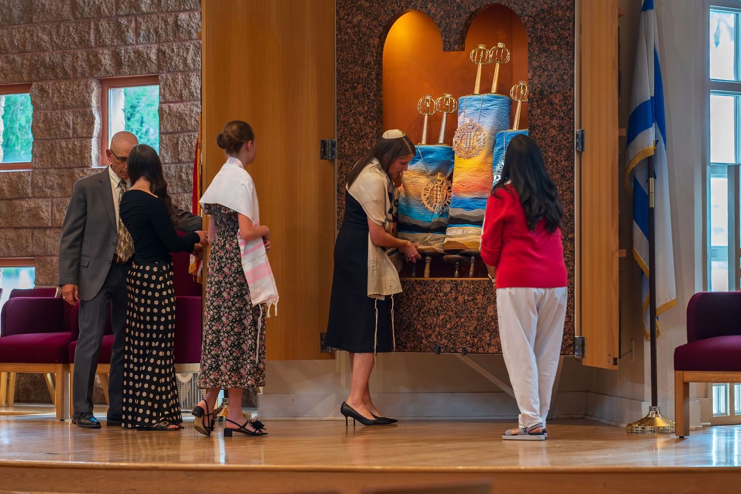 People gather around colorful artwork displayed on a stage in a synagogue with Israeli flag visible.