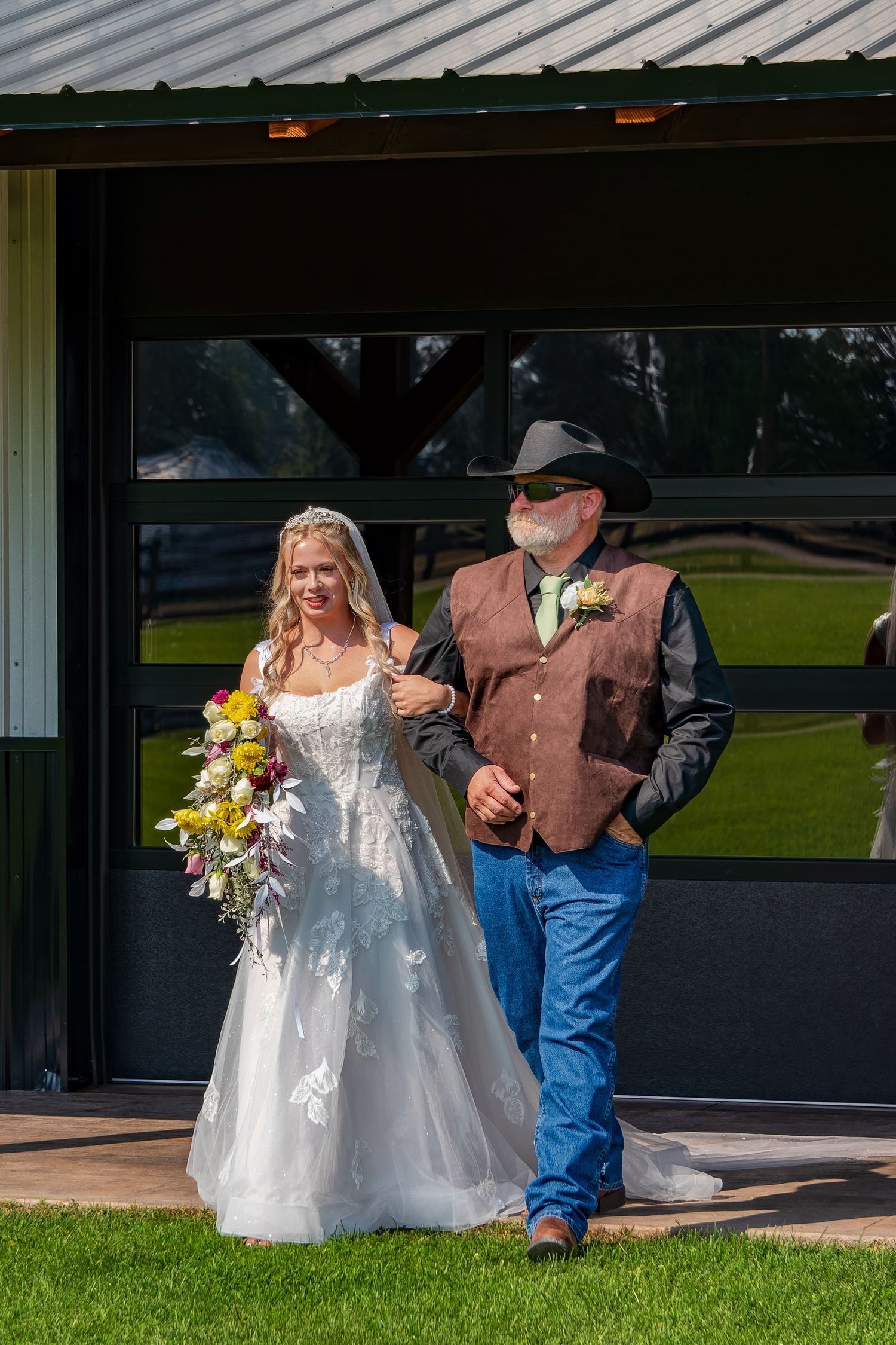 A bride in a white dress walks with someone wearing jeans and a brown vest during an outdoor wedding ceremony.