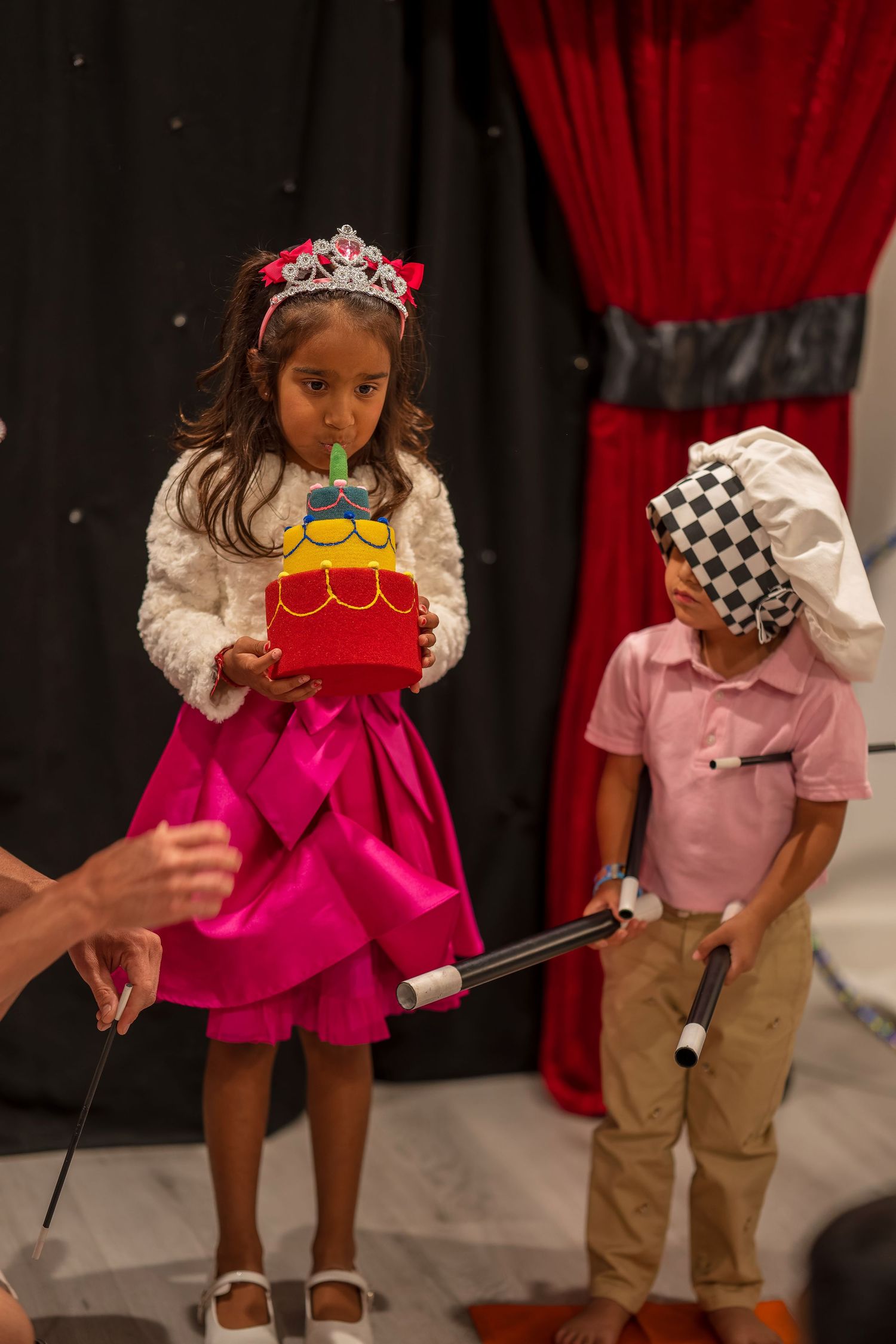 Child in a festive pink dress and tiara participates in a magic trick on stage.