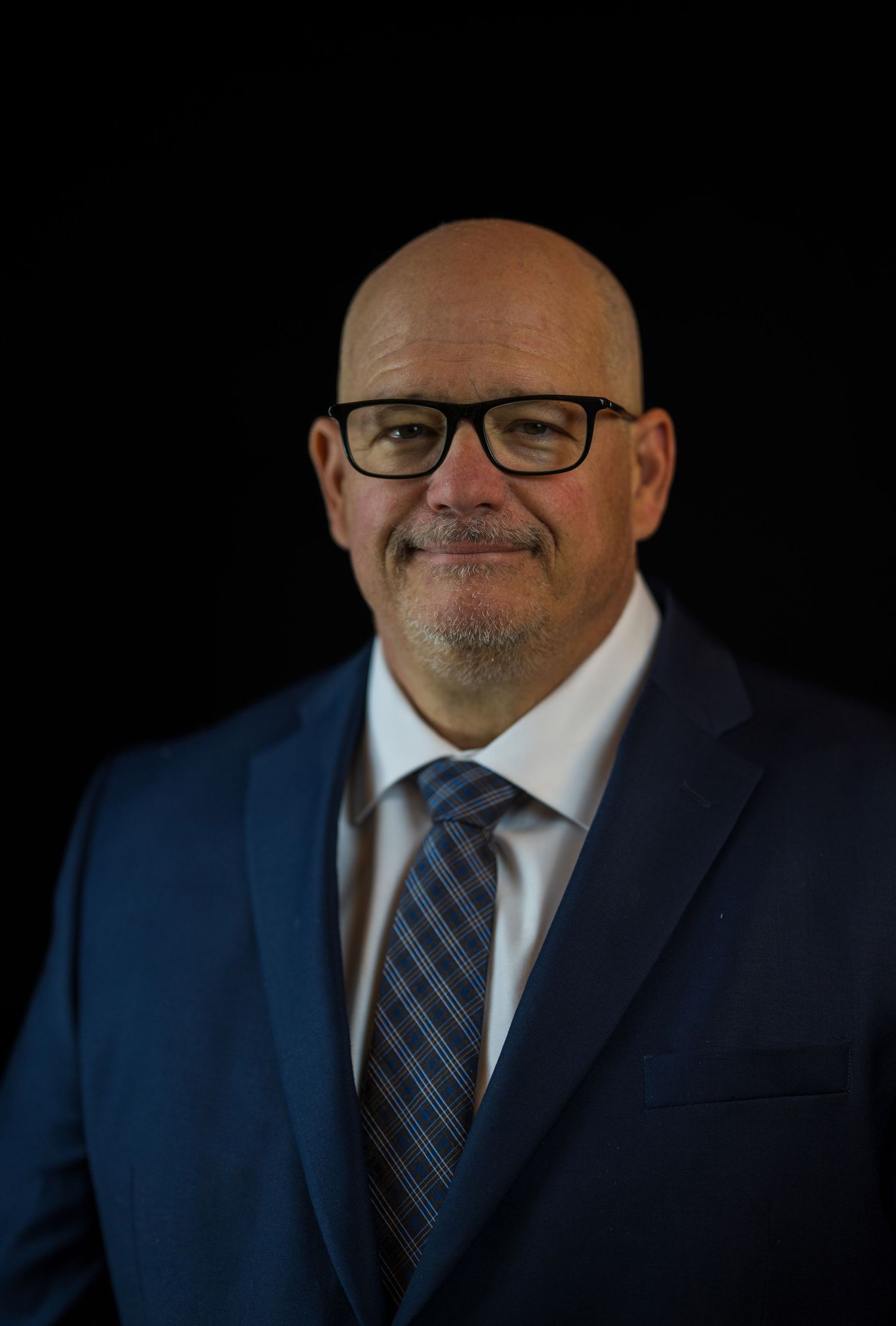 Professional headshot of a person in a navy suit and striped tie against a dark background.