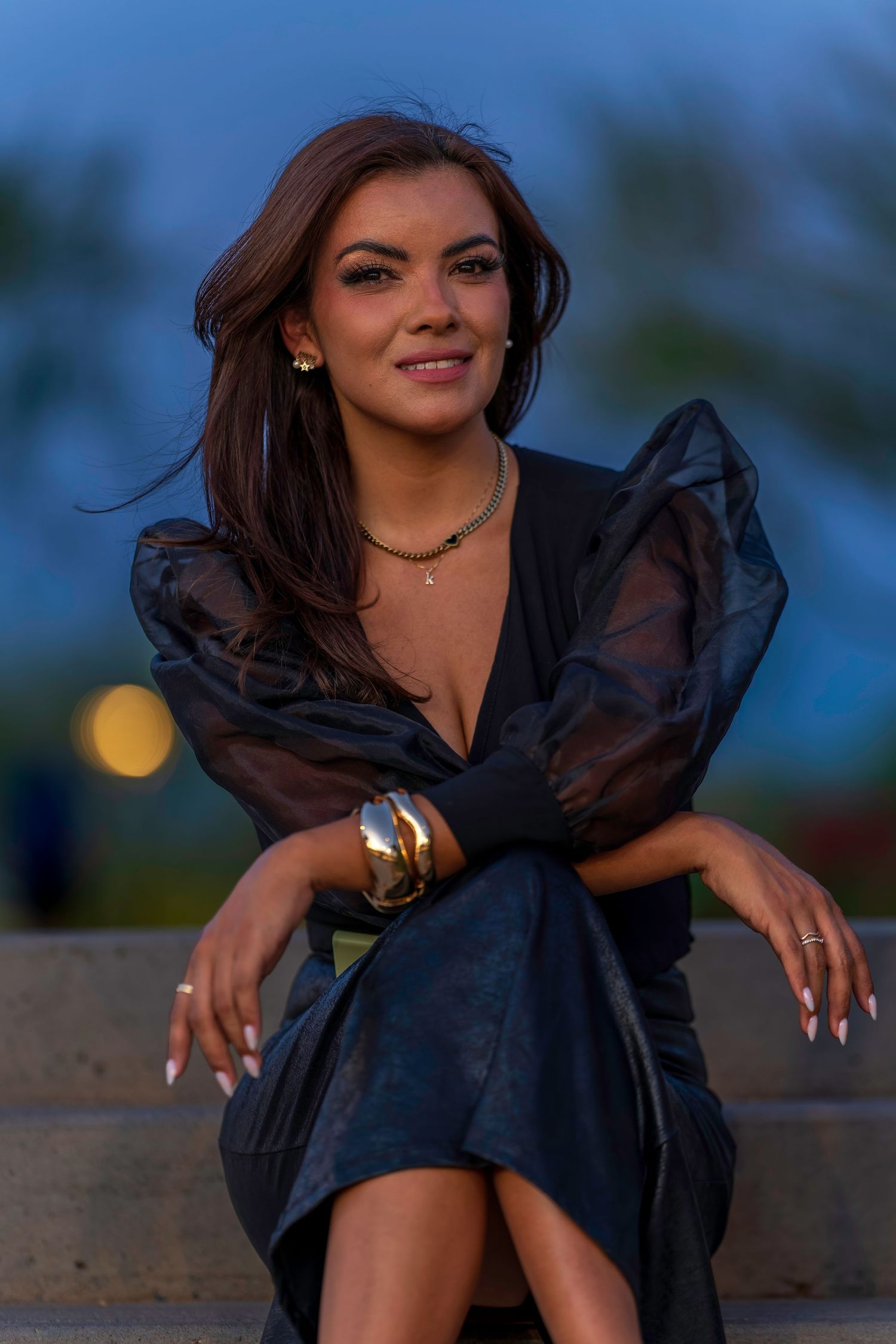 Smiling brunette woman in black sheer puff-sleeve dress sitting on steps at dusk.