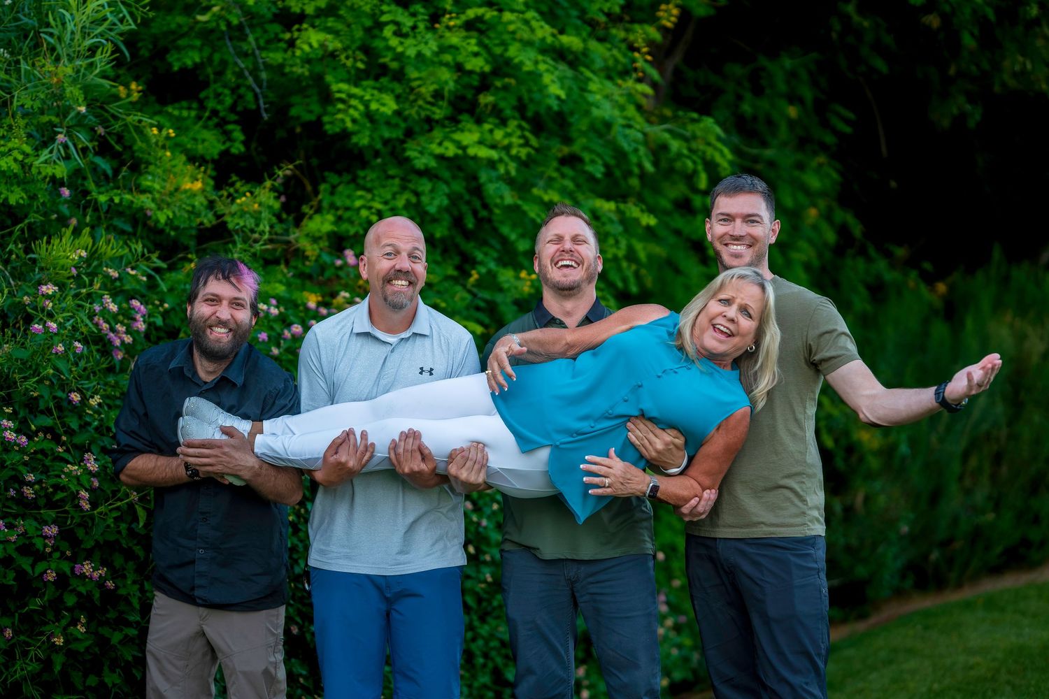 Four men joyfully lift a laughing woman horizontally in a lush green garden setting.