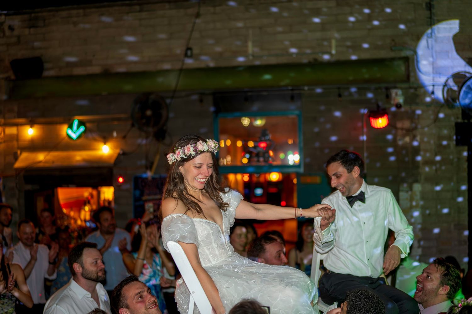 Bride lifted by guests during joyful wedding reception dance celebration in a warmly lit venue.