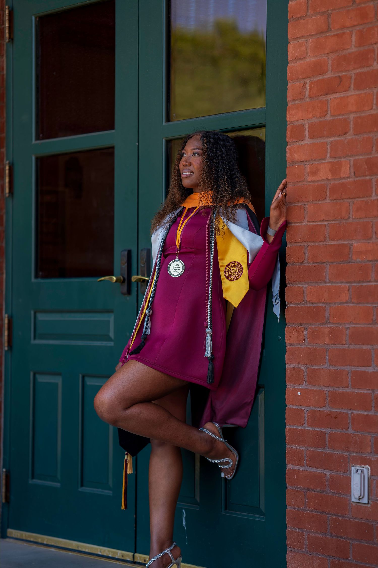Young Black woman in maroon graduation gown and stoles poses by green doors of brick building.