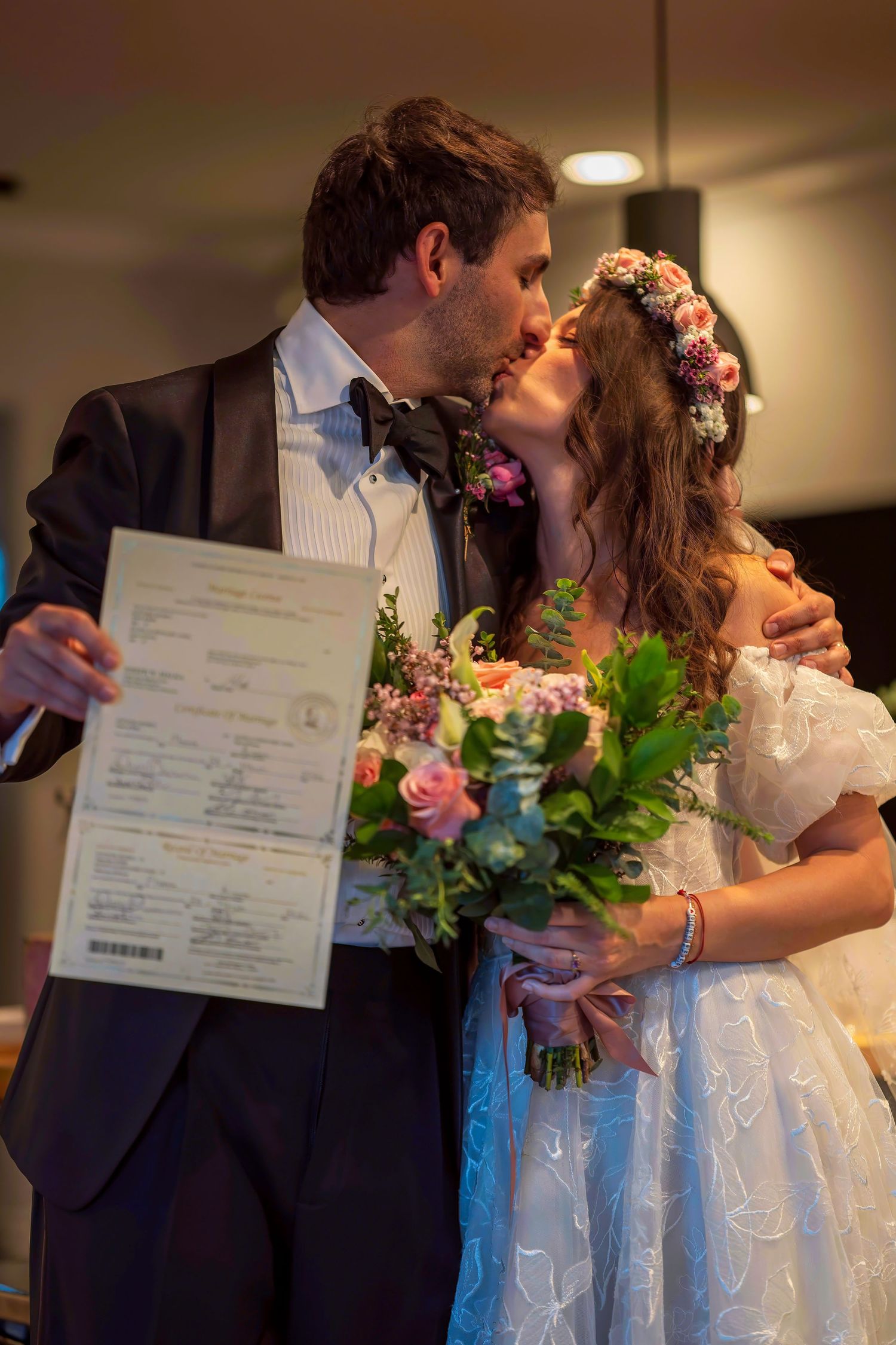 Newlyweds sharing a kiss while holding marriage certificate and bridal bouquet indoors.