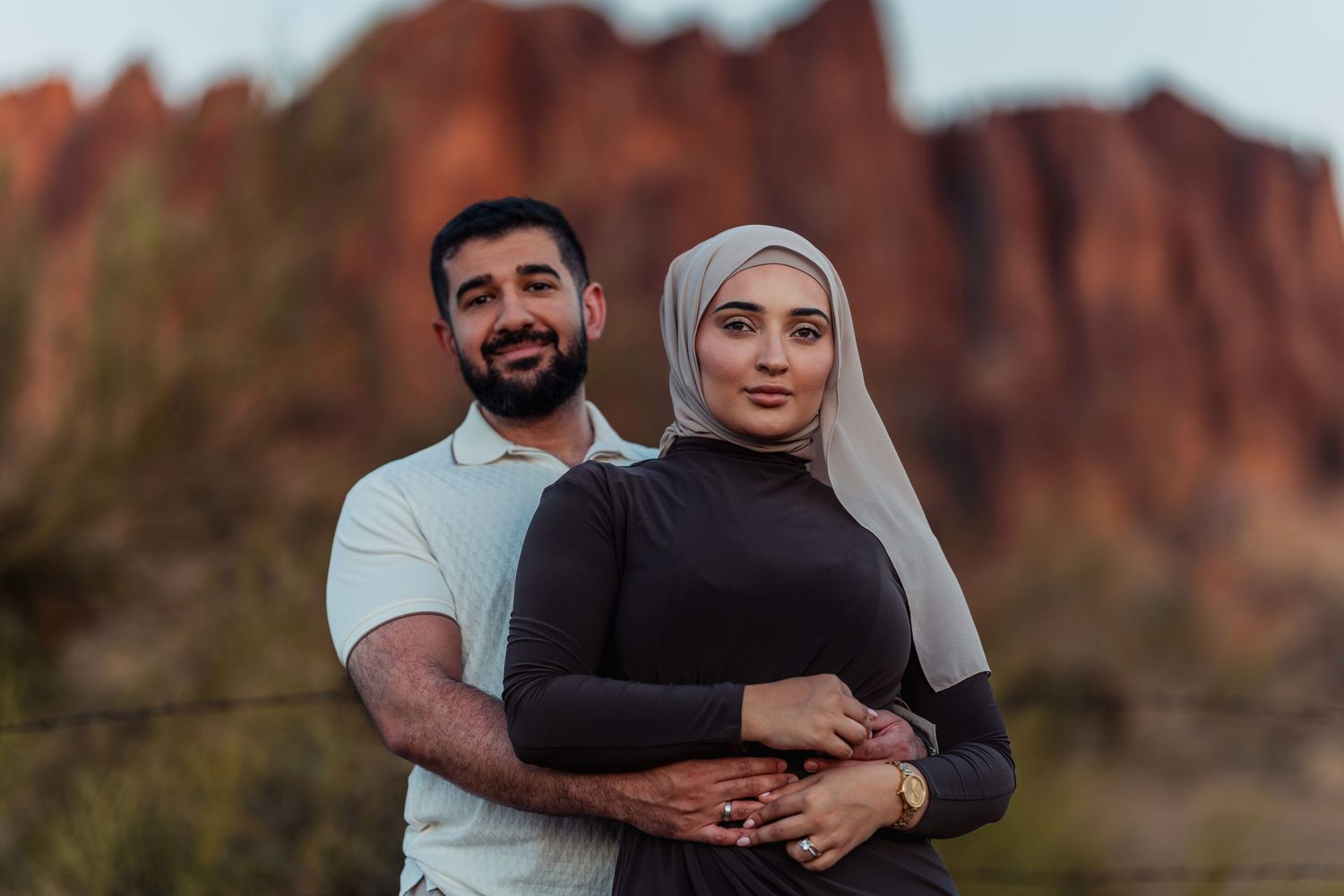 Muslim couple posing together at red rock canyon, woman in hijab and black dress with man in white polo embracing warmly.