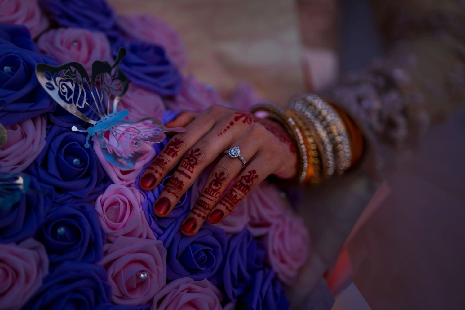 Bride's hand with henna, diamond ring, and gold bangles resting on purple and pink roses bouquet.