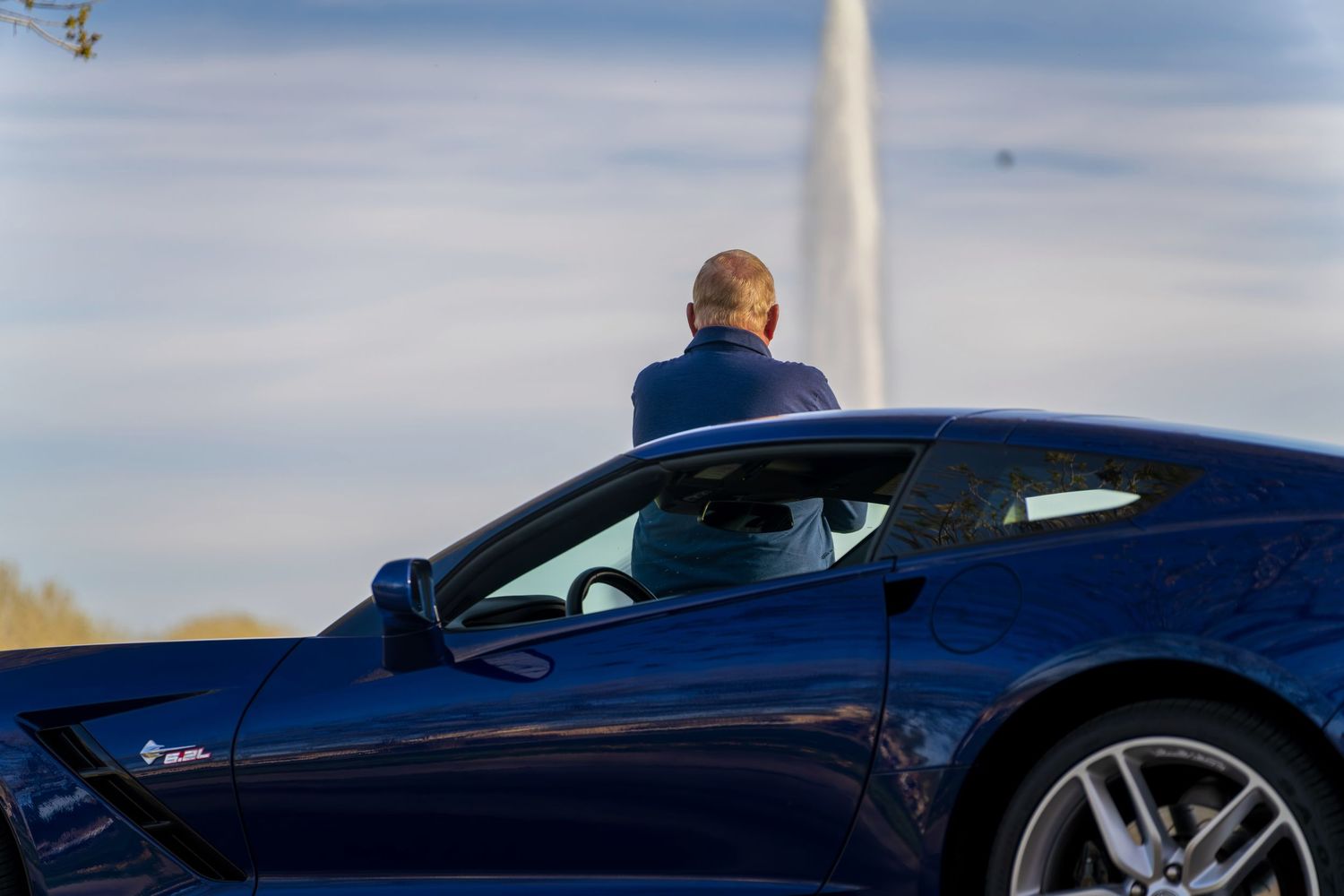 A man standing beside a sleek dark blue sports car, possibly a Corvette, under a partly cloudy sky.