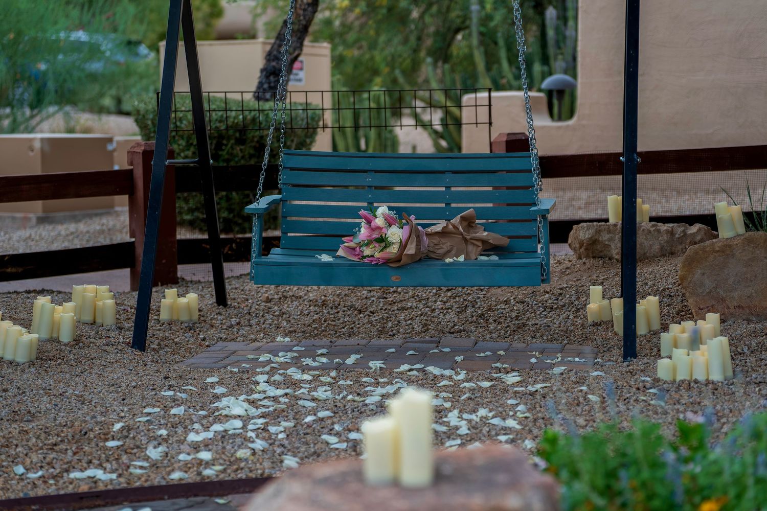 A dog rests on a teal porch swing surrounded by lit candles in a desert garden setting.
