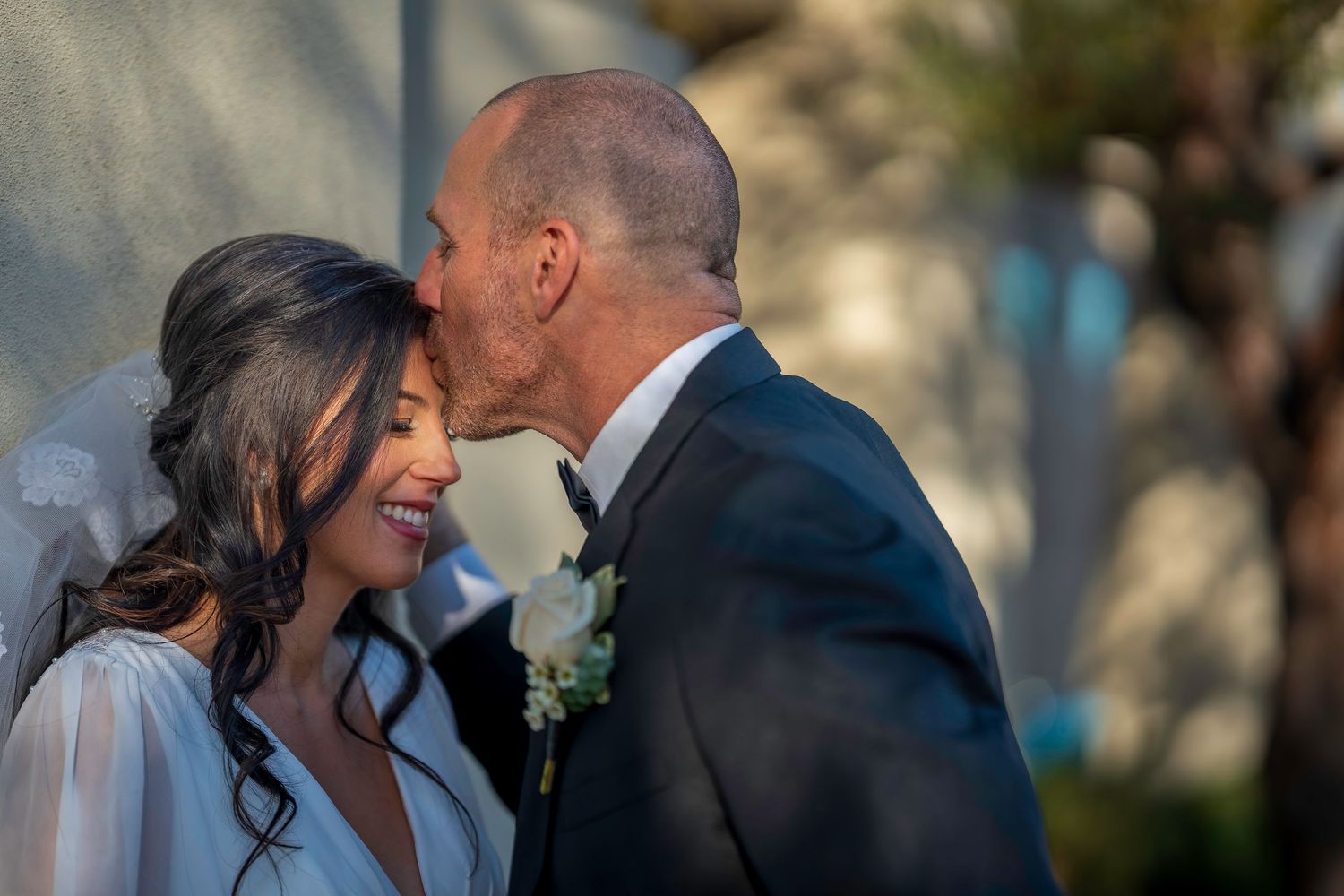 Groom gently kisses smiling bride on the cheek during outdoor wedding ceremony near white floral decor.
