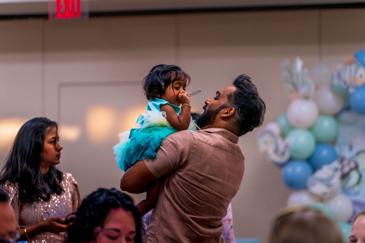 Father lovingly holds up his toddler daughter in teal dress at a festive celebration with balloon decorations.