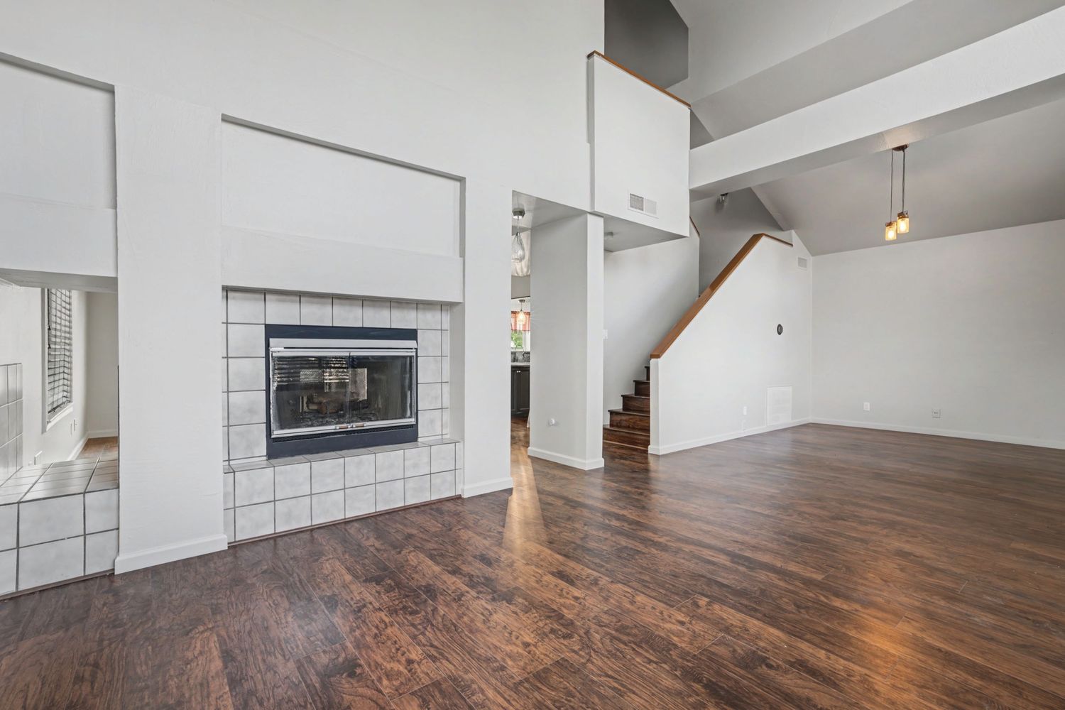 Spacious living room with dark hardwood floors, white walls, tile fireplace, high ceilings, and staircase in background.