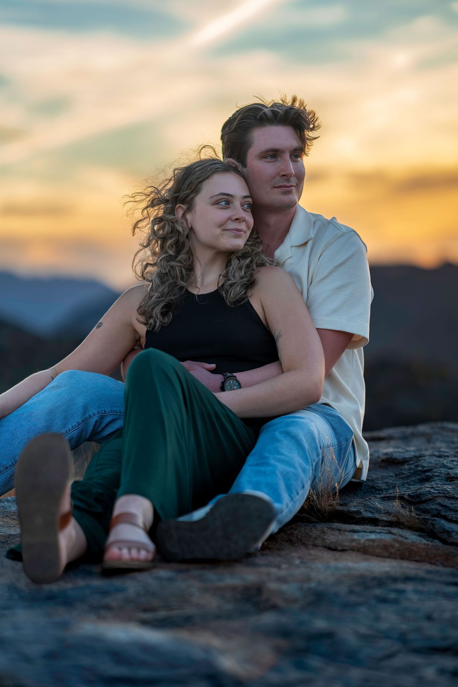 Couple sitting together on rocky surface watching a vibrant orange and yellow sunset over mountain landscape.